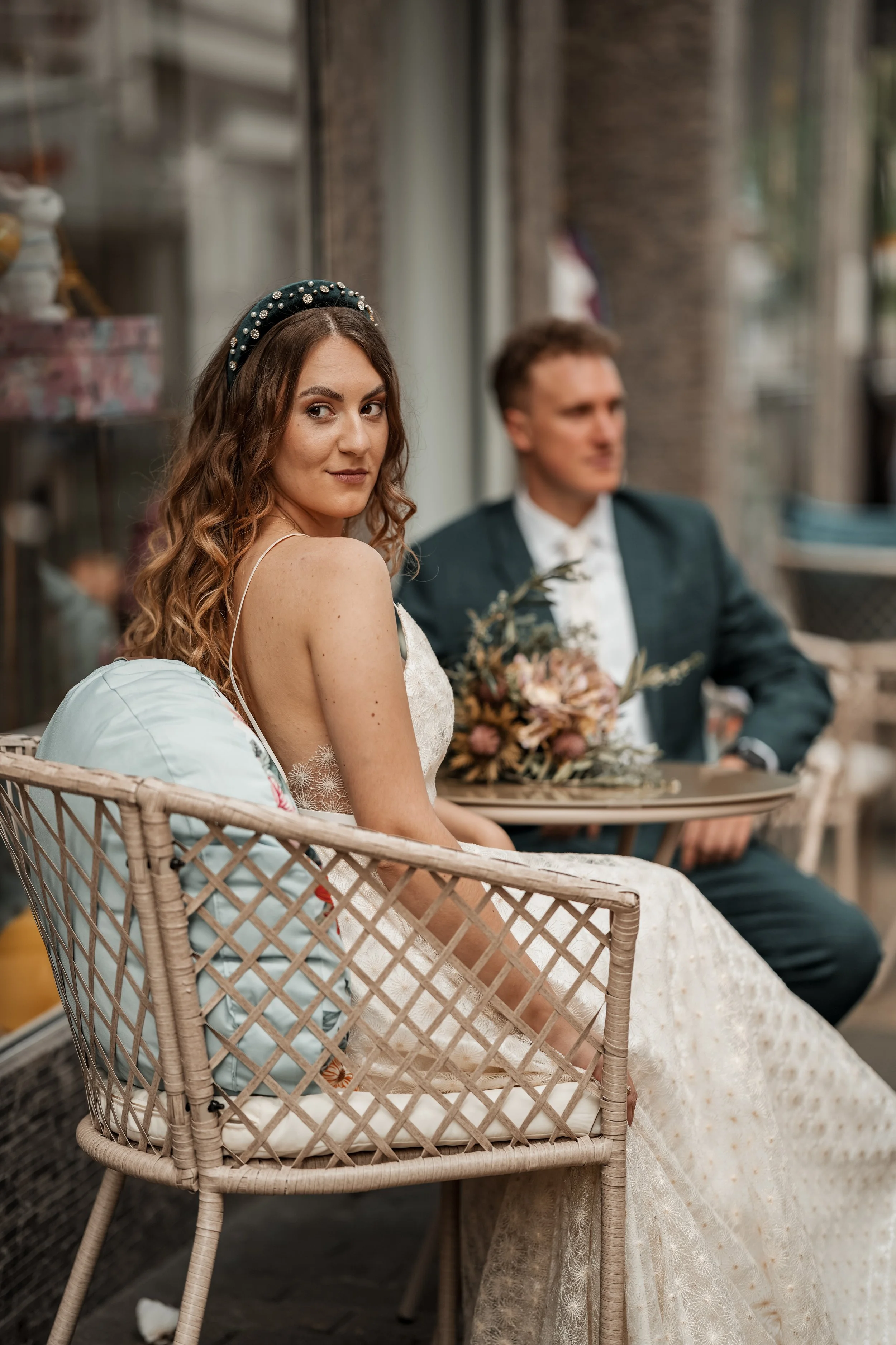 Eine Braut in einem weißen Kleid sitzt auf einem Gartenmöbel mit einem Mann im dunklen Anzug im Hintergrund, der eine Hochzeitsdekoration hält.
