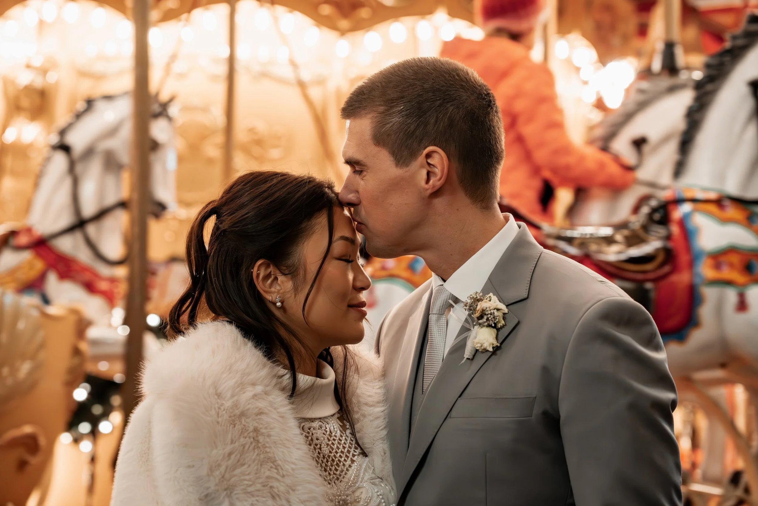 Ein Brautpaar steht sich in einer romantischen Umarmung und küsst sich, vor einem Karussell bei einem festlichen Anlass.