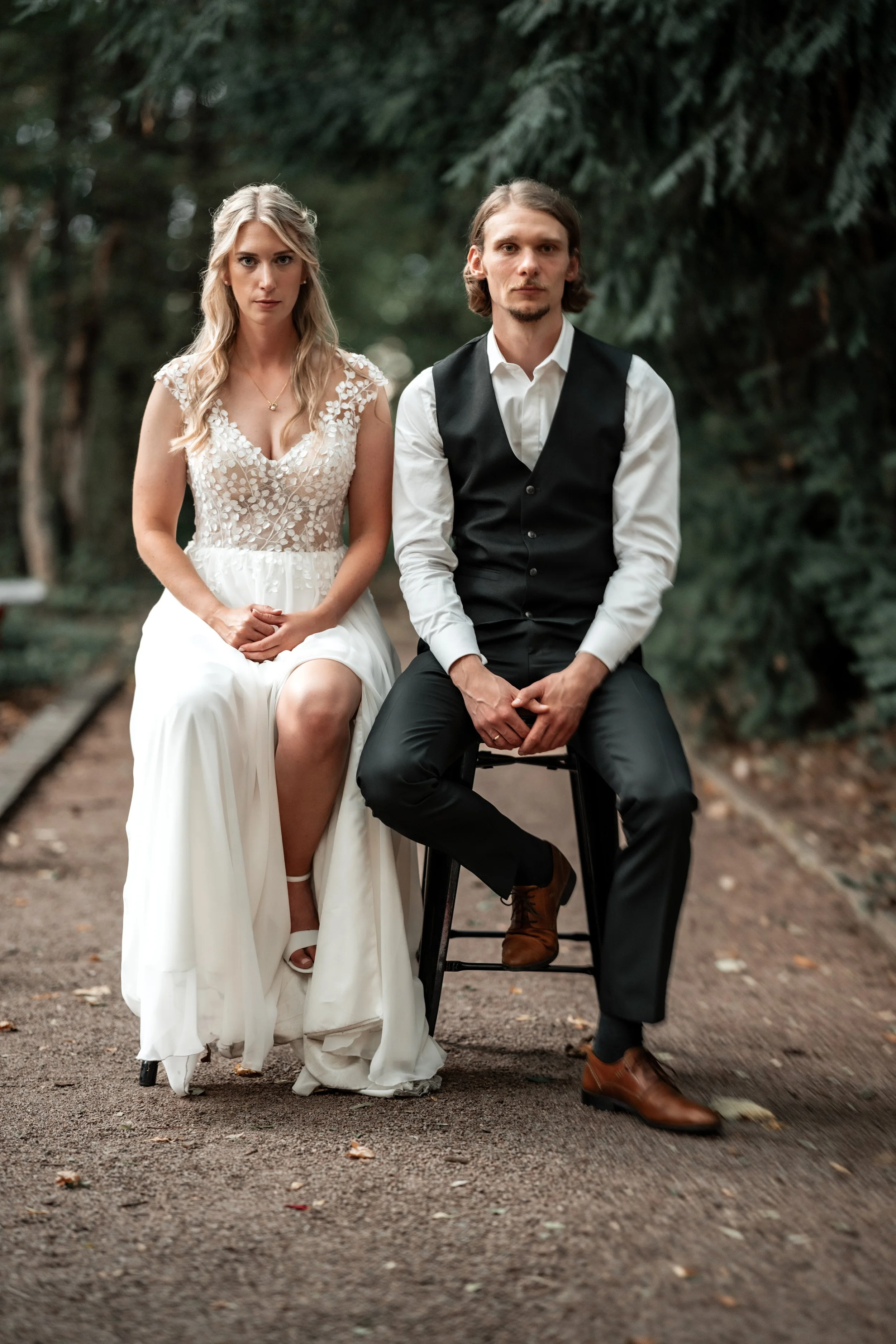 Ein Brautpaar sitzt auf einem Baumstamm in einem Wald, die Frau trägt ein weißes Brautkleid und der Mann trägt eine schwarze Weste mit weißen Hemd, beide schauen ernst in die Kamera.