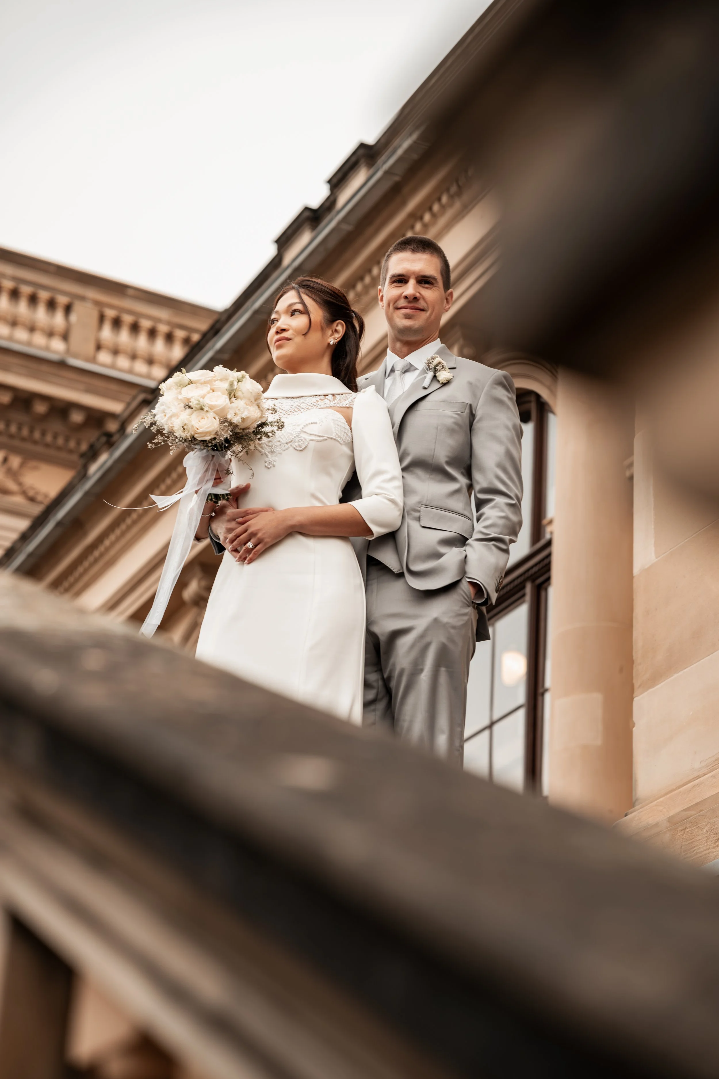 Ein Brautpaar steht auf einer Treppe vor einem historischen Gebäude, die Braut hält einen Blumenstrauß, der Bräutigam trägt einen grauen Anzug.
