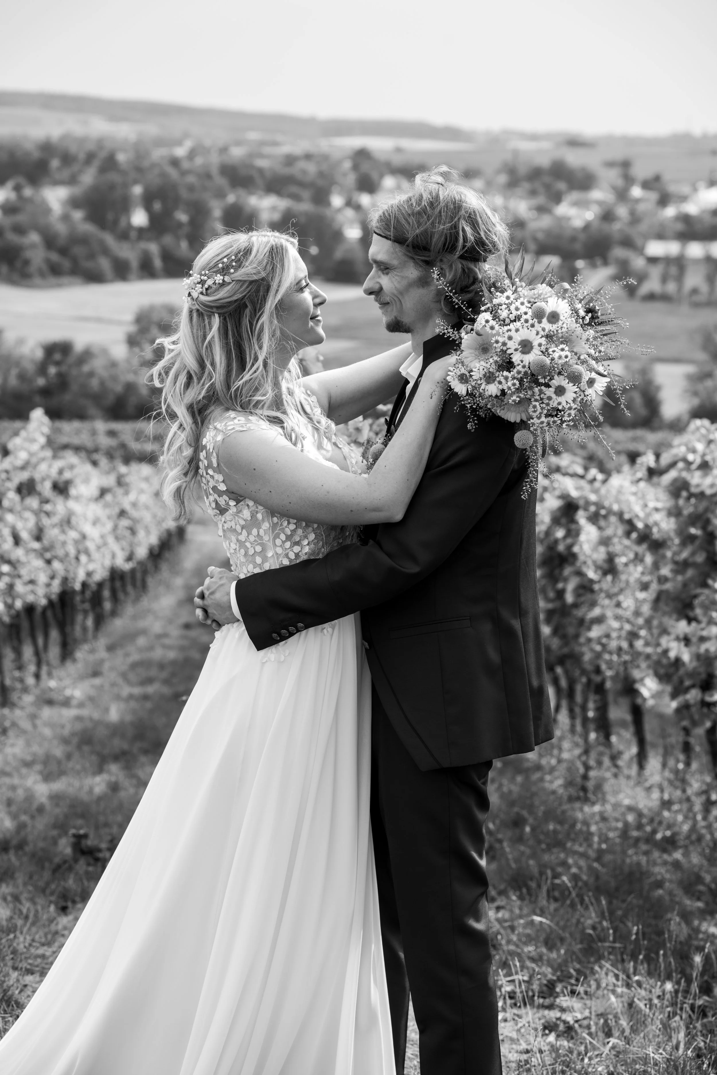 Braut und Bräutigam in einer ländlichen Weinberglandschaft, in die sich verliebt ansehen, mit Blumenstrauß und Hochzeitskleidung, im Schwarz-Weiß-Format.