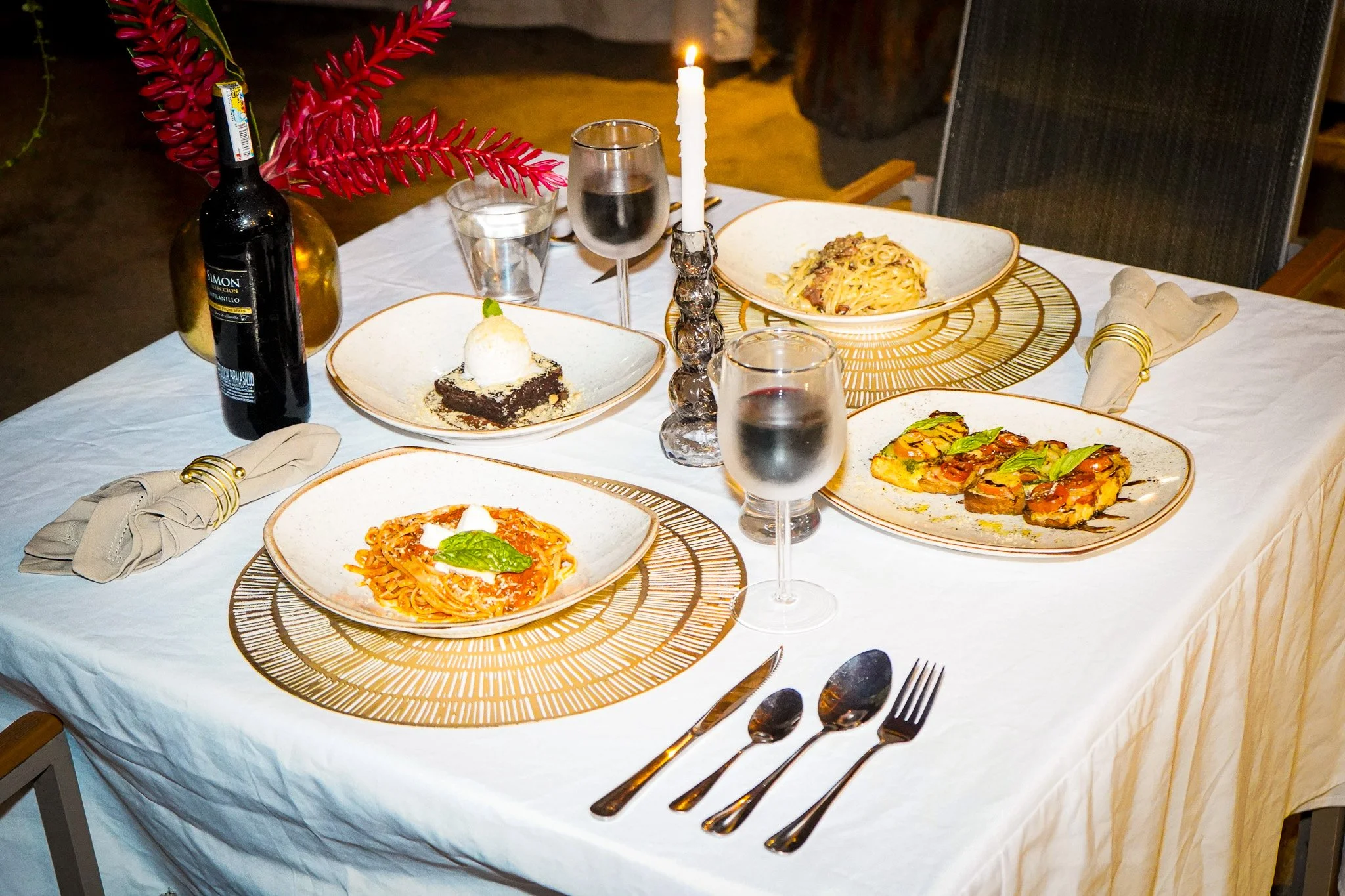 A dinner table set for a meal with plates of pasta, a slice of dessert, wine glasses, a bottle of red wine, a candle, and a decorative flower arrangement.