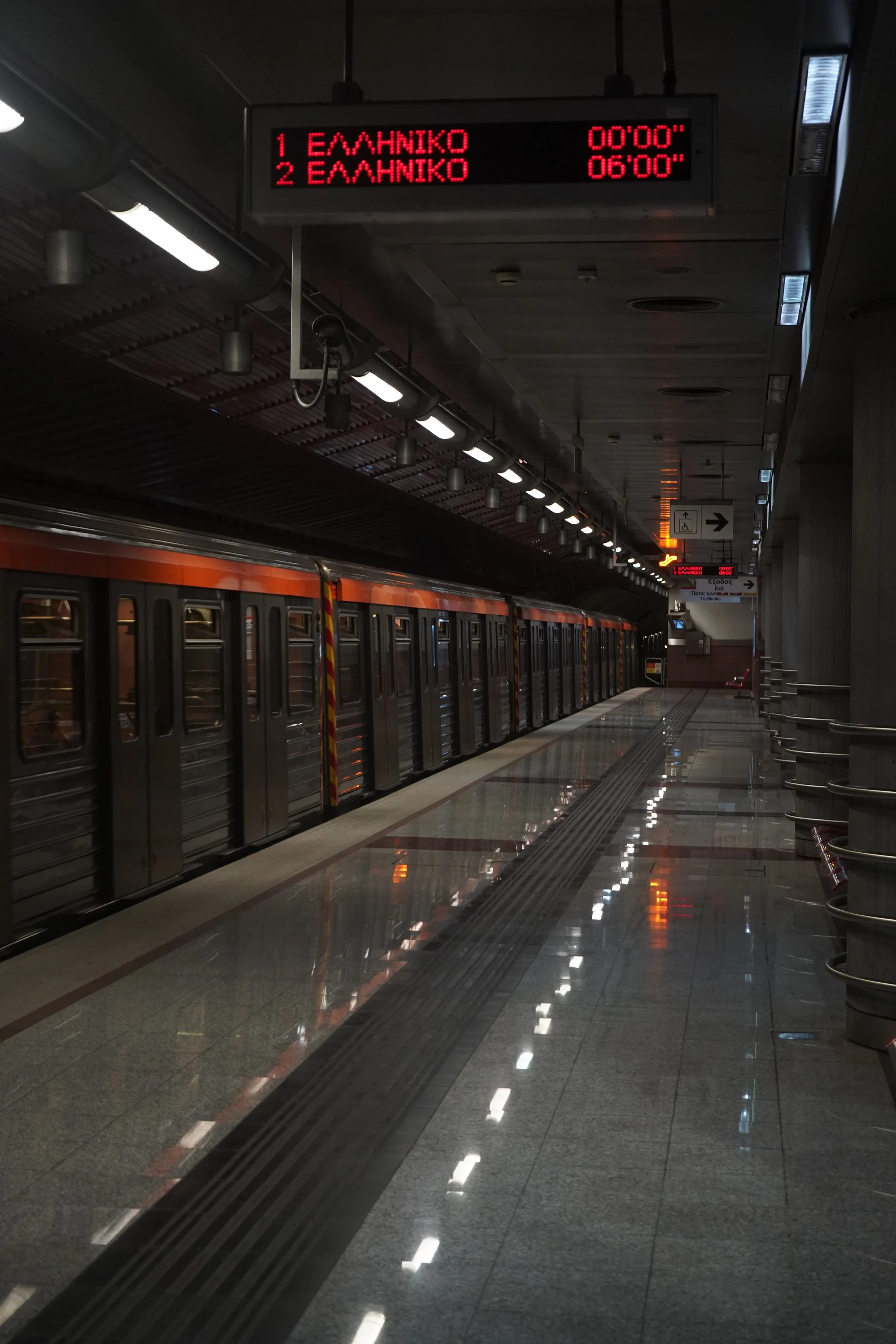 Empty subway platform with a train stopped at the station. Overhead electronic display board showing train departure times and destinations written in Greek.
