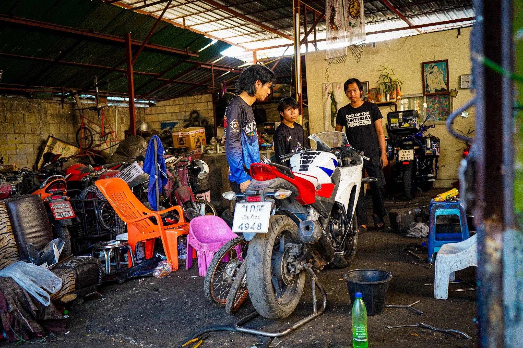 Three children inside a cluttered workshop with motorcycles, chairs, and various items. One child is looking at a motorcycle, and the others stand nearby. The background shows shelves, pictures, and tools.