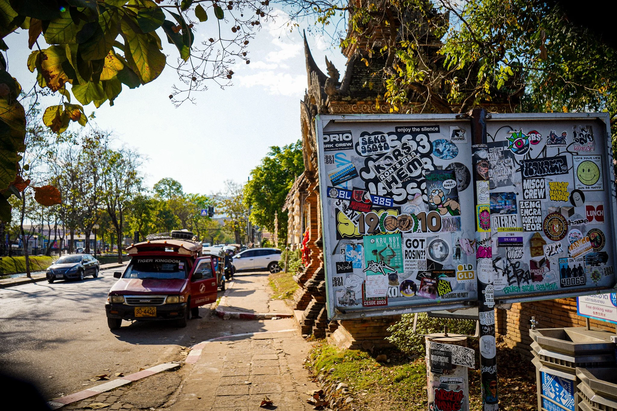 A busy street scene with cars driving past, a sidewalk with advertisements and stickers, and a traditional temple or building partially visible in the background surrounded by trees.