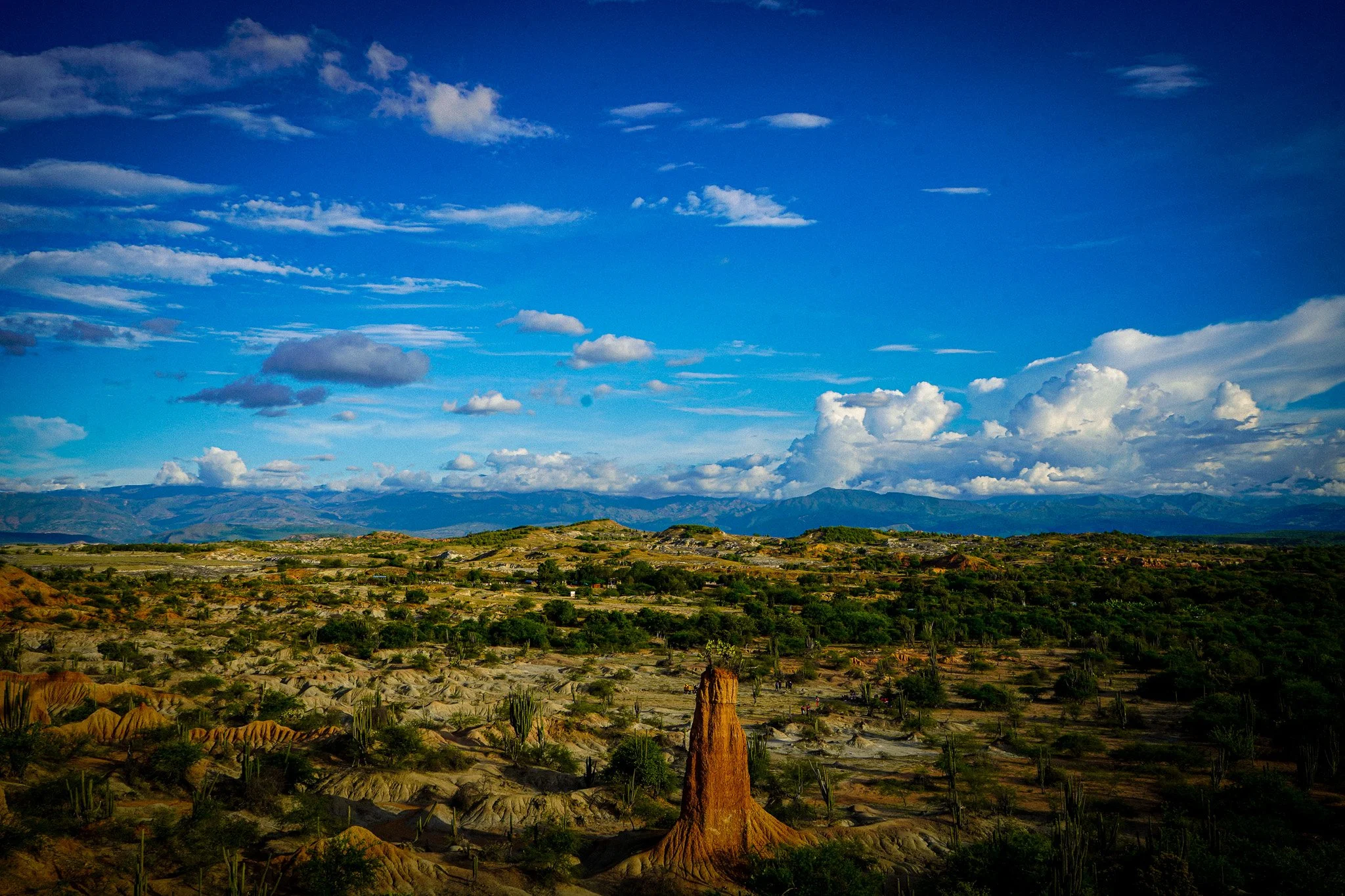 Scenic landscape of a semi-arid area with sparse vegetation, unique rock formations, and distant mountains under a bright blue sky with scattered clouds.