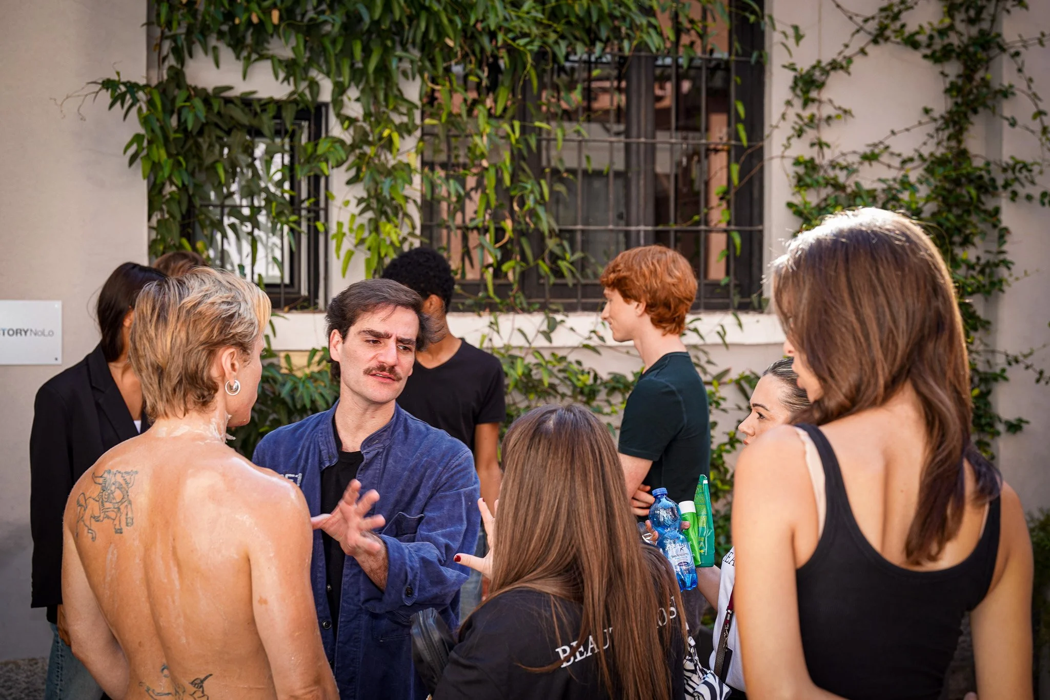 A group of people gathered outdoors, engaging in conversation. Some are women with varying hair colors and styles, and a man with dark hair and a mustache. One person with tattoos and bare back is also present. The background features a building with