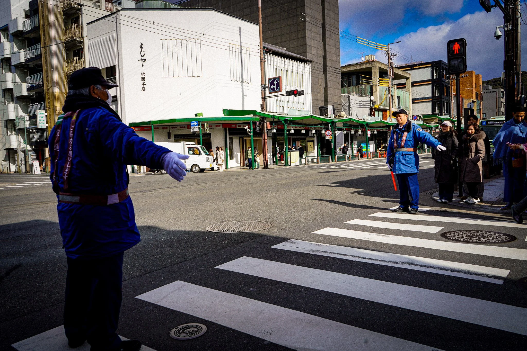Pedestrians crossing street at a crosswalk with a traffic light showing red hand signal in an urban area with buildings, utility poles, and a bus stop shelter.