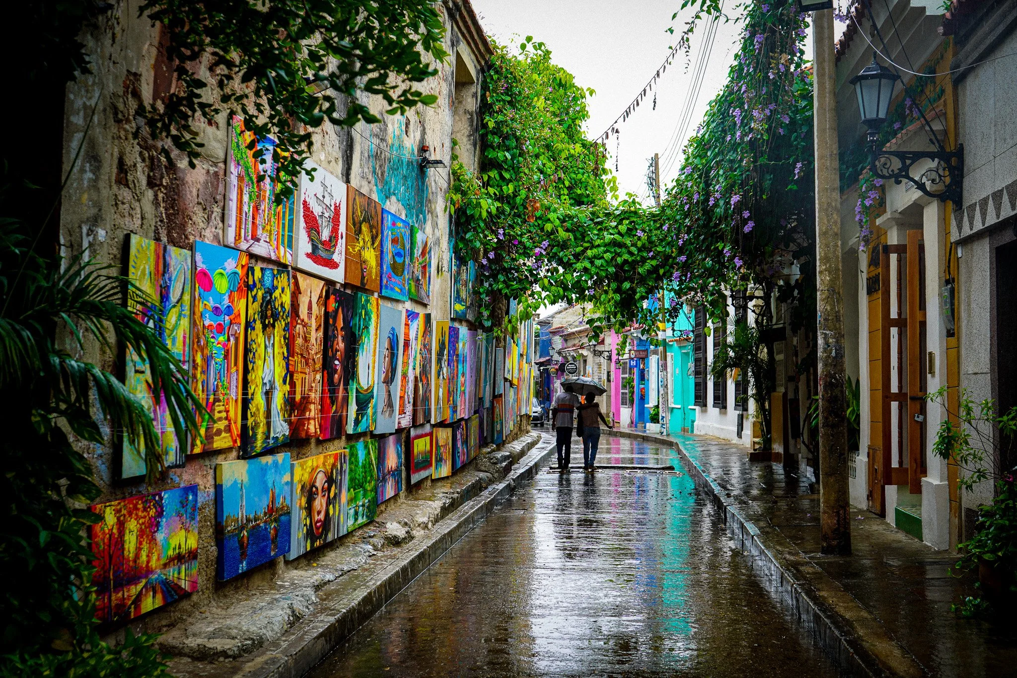 A rainy street with colorful paintings on display along a stone wall on the left and brightly painted buildings on the right. Two people walk under umbrellas, and lush greenery and flowering plants hang overhead.
