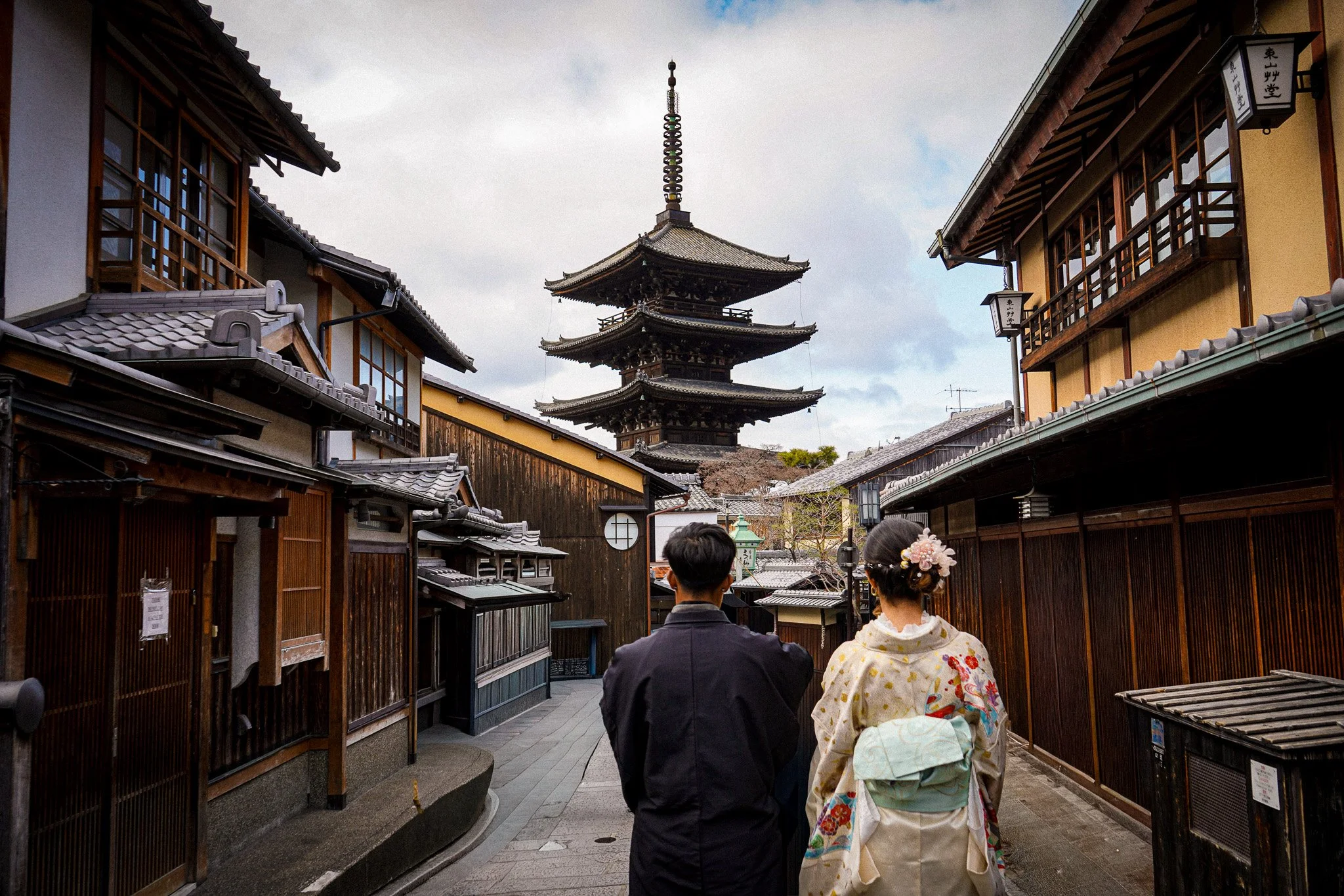 A traditional Japanese street scene with a pagoda in the background and two people wearing kimono walking away from the camera.