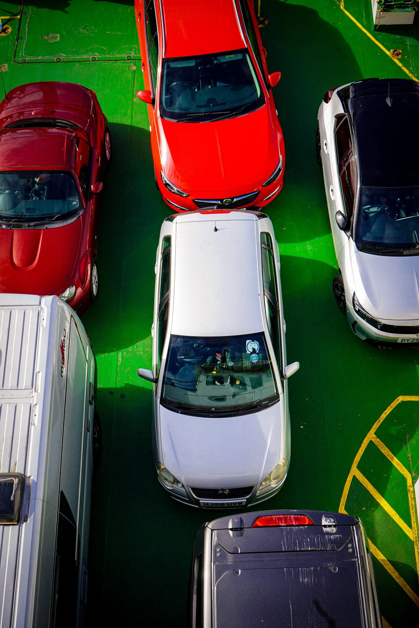 A top-down view of a parking lot with several parked cars on a green painted surface, including a red car in the center, surrounded by other red, white, black, and gray vehicles.