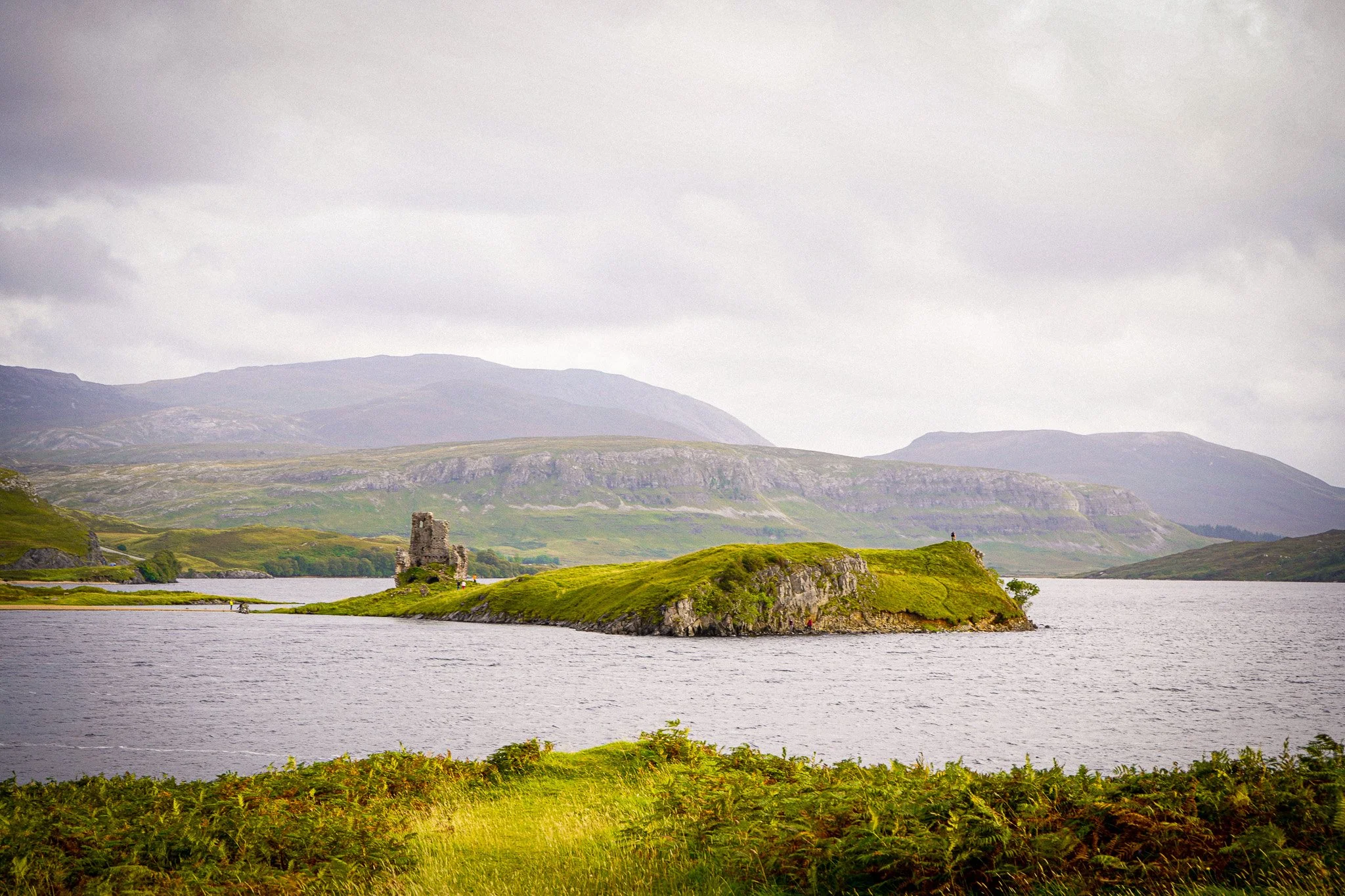 Scenic view of a lake with green grassy islands and ruins of an old stone structure, mountains in the background, overcast sky.