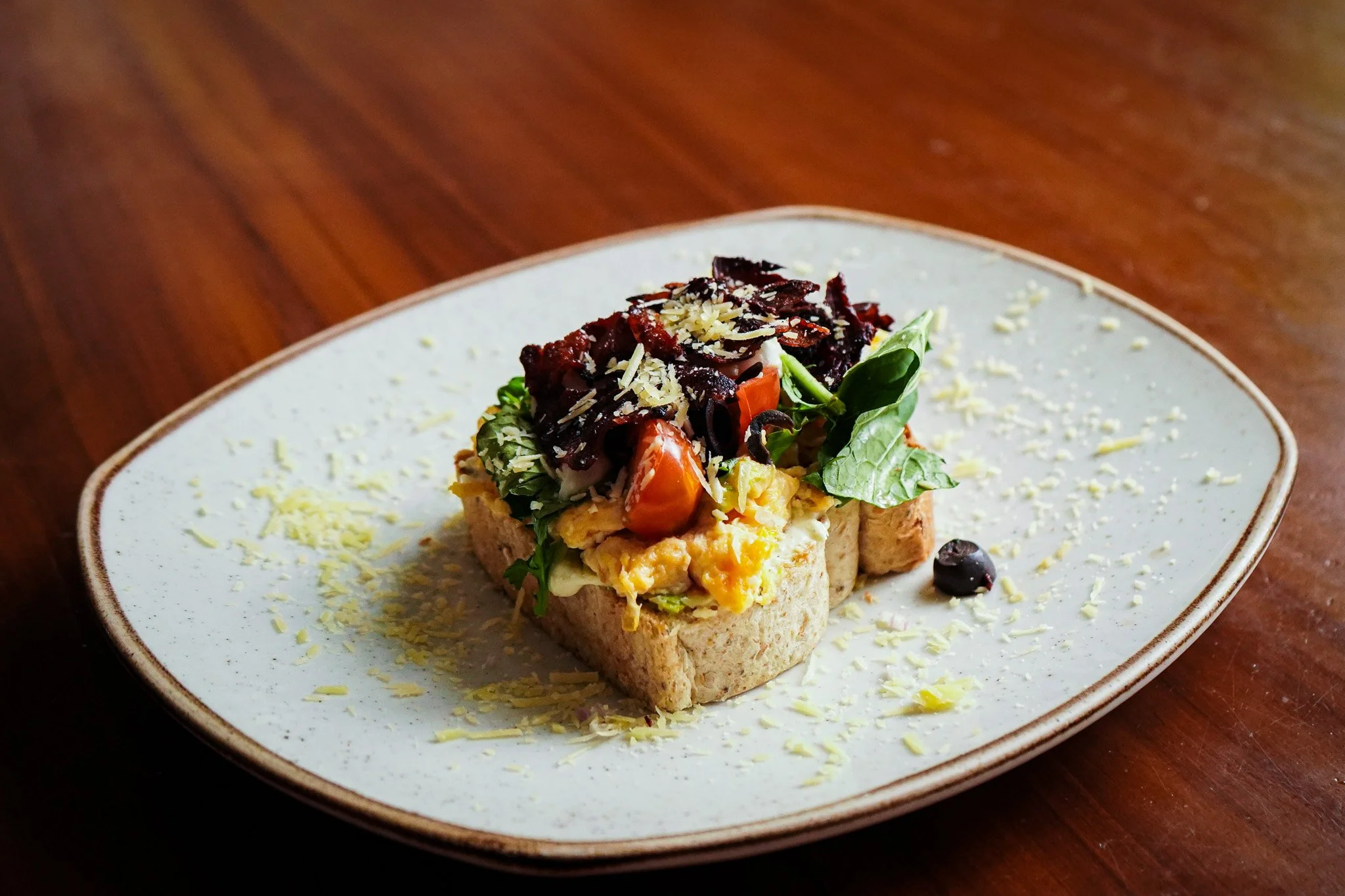 A plate with an open-faced sandwich featuring scrambled eggs, vegetables, and shredded cheese, garnished with basil leaves and grated cheese on a ceramic plate.