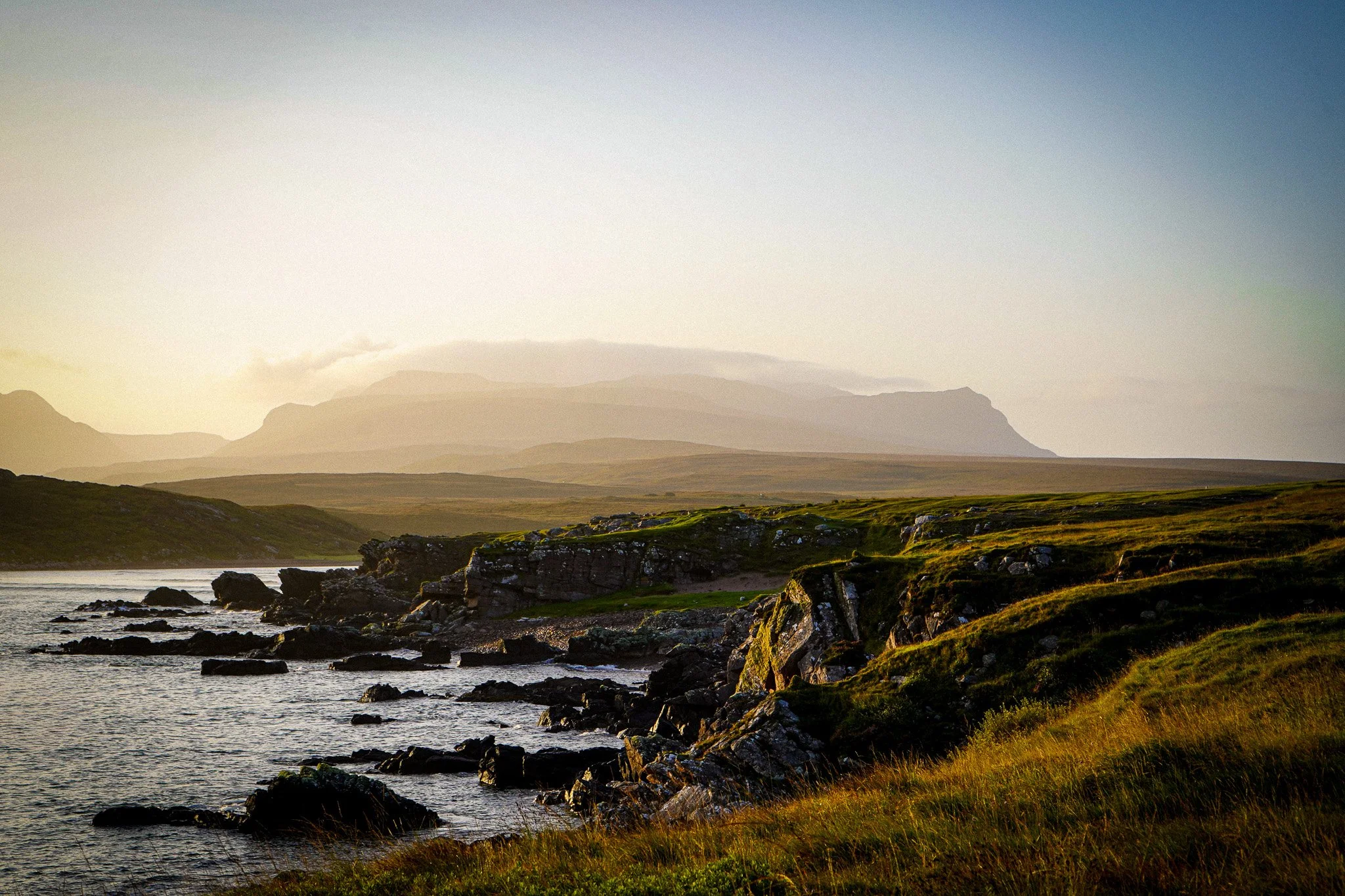 Scenic landscape of a coastline with rocky shoreline, green grass, and rolling hills under a clear sky at sunset.