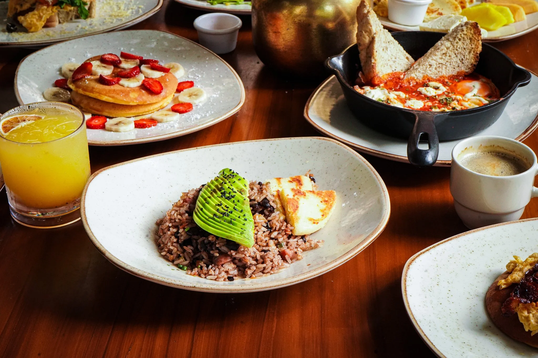 A breakfast spread featuring a plate of sliced strawberries and bananas on pancakes, a glass of orange juice, a bowl of rice with avocado slices, a skillet with fried eggs topped with toast, and a cup of coffee served on a wooden table.