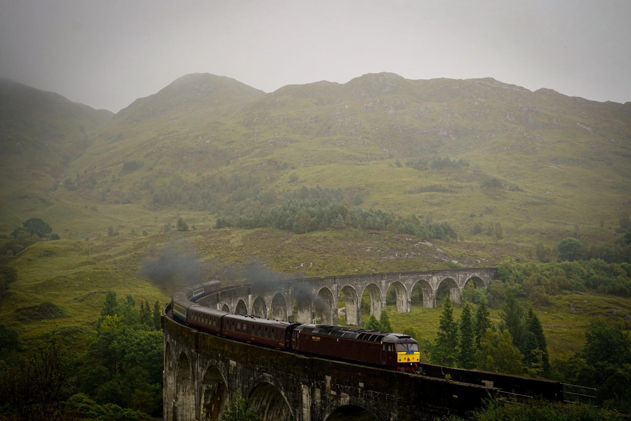 A vintage train traveling across a stone arch bridge in a lush green valley with mountains in the background under an overcast sky.