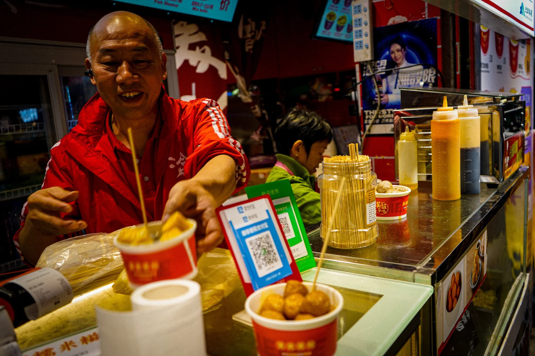 A man in a red shirt with white stripes smiling behind a counter at a food stand with condiments and cups of food, with two women in the background and digital menu screens above.