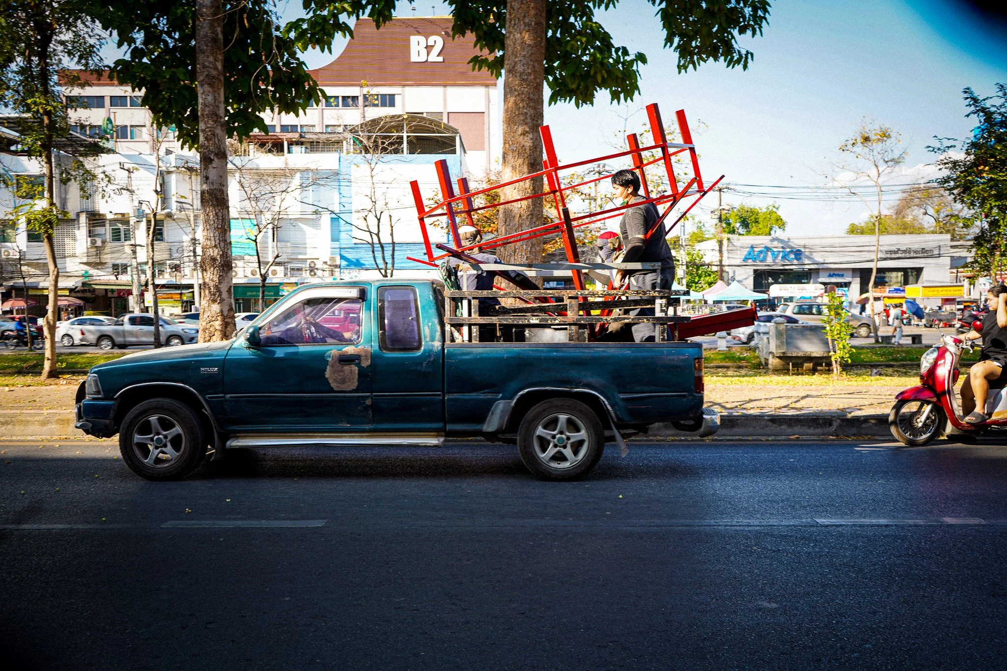 A man in gray clothing and a face mask is standing on a flatbed truck, setting up or adjusting red metal frames that resemble chairs or stools, parked on a street with several cars and motorcycles. Behind them is a busy urban area with trees, buildin