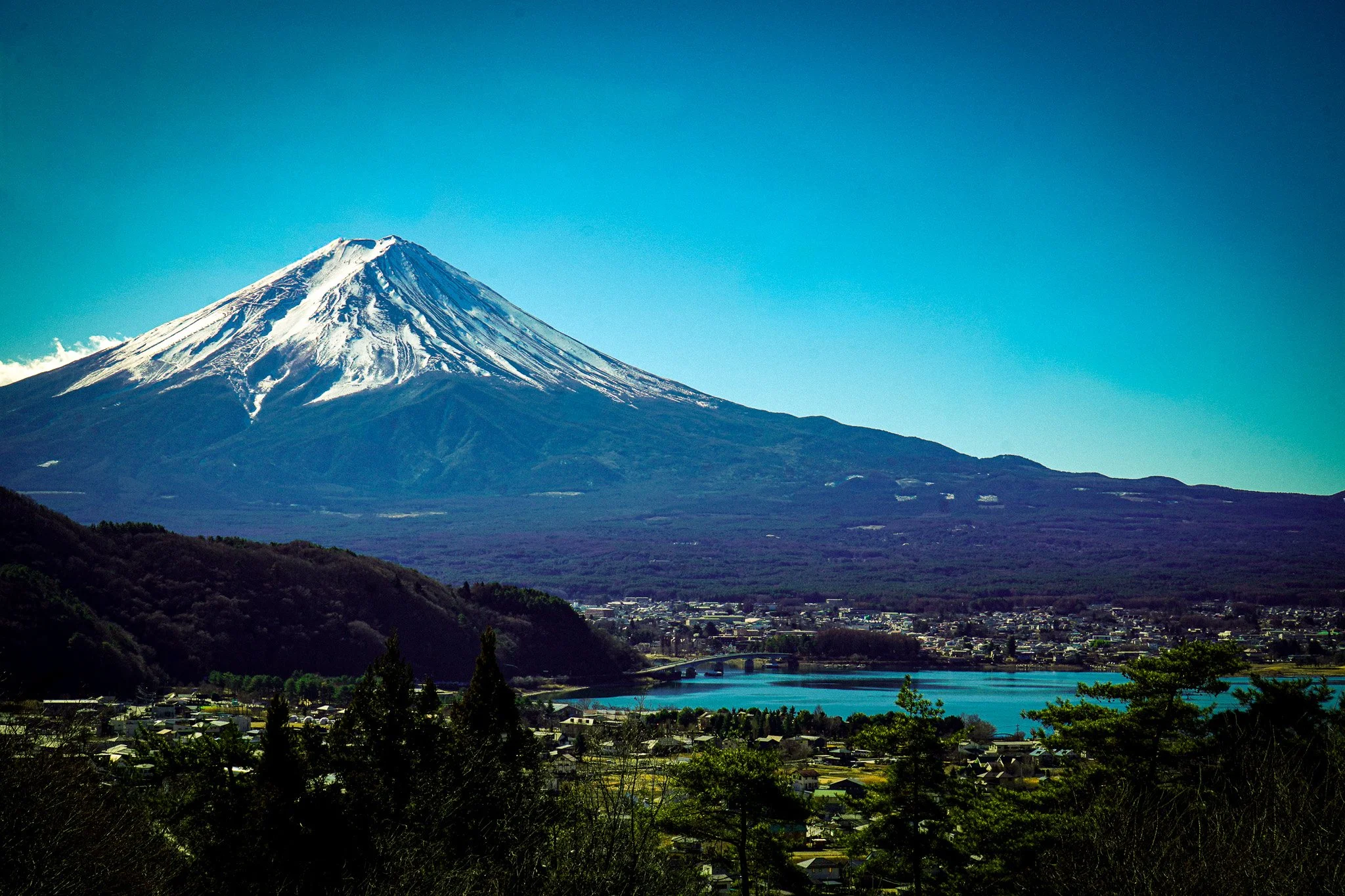 Snow-capped Mount Fuji with a city and river in the foreground under a clear blue sky.