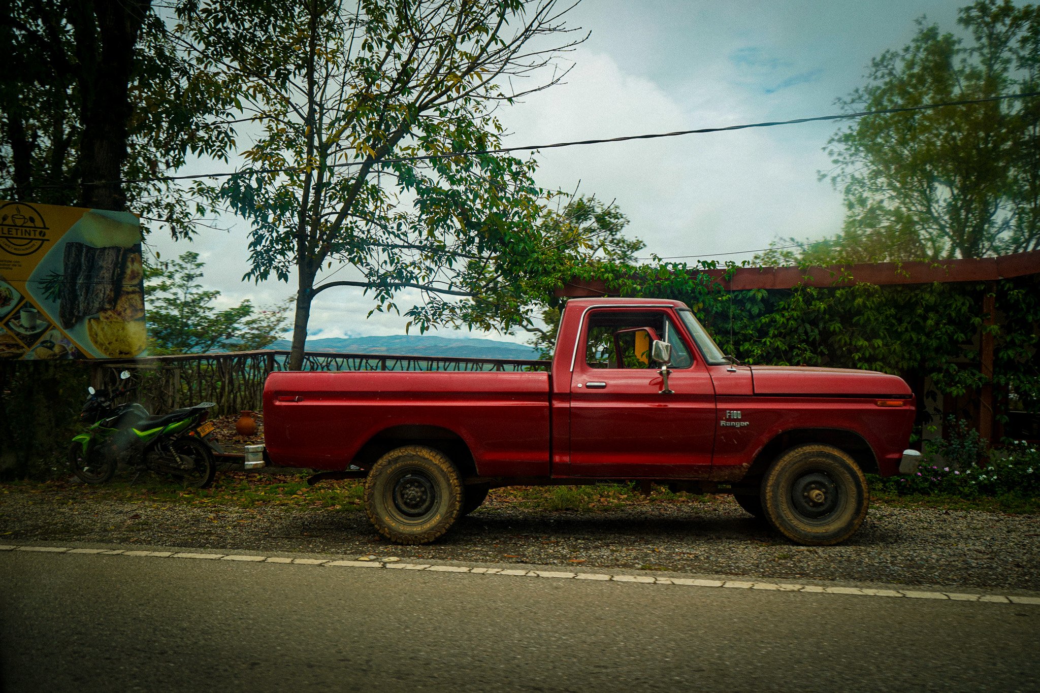 A red pickup truck parked on the side of a road with trees and a cloudy sky in the background. There are motorcycles parked nearby and a partially visible billboard on the left side.