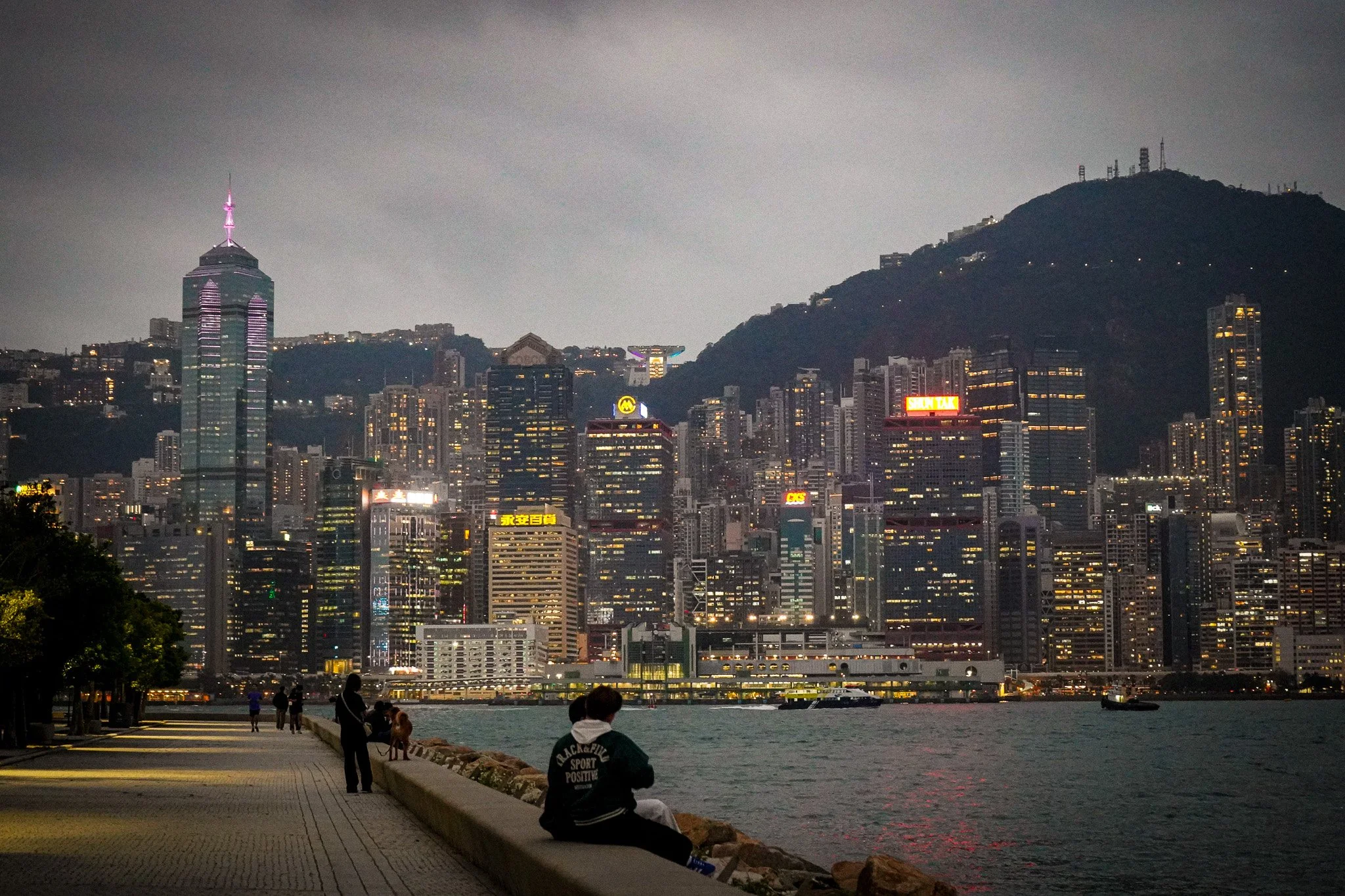 Nighttime city skyline with tall buildings, including the IFC Tower, and a hill with communications towers. People are walking and sitting along a waterfront promenade.