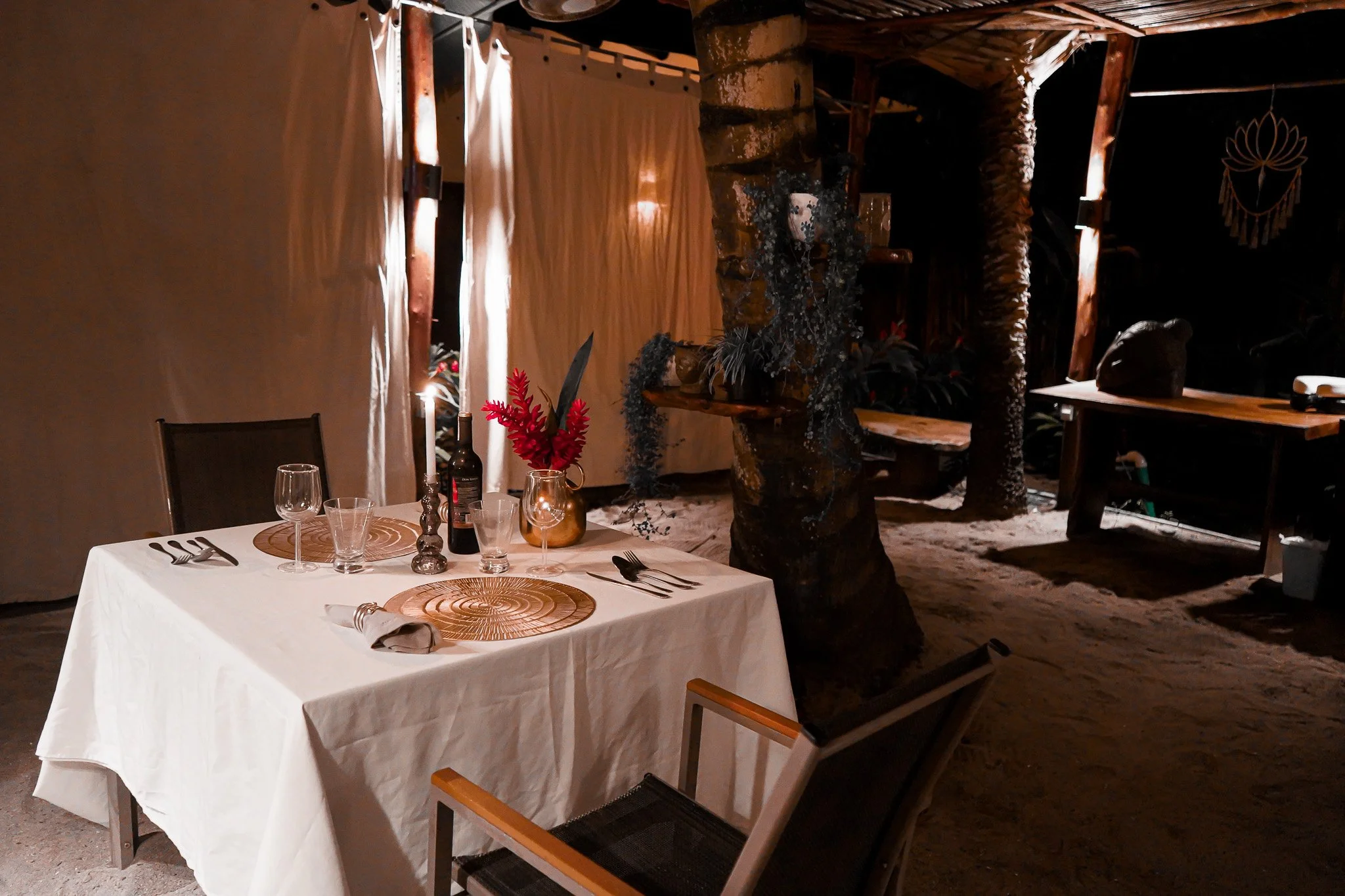 A dining table set for a meal with a white tablecloth, wine glasses, cutlery, and decorative wooden placemats. There is a small bottle with red flowers on the table. The background shows a rustic setting with a tree trunk and wooden beams, and a curt