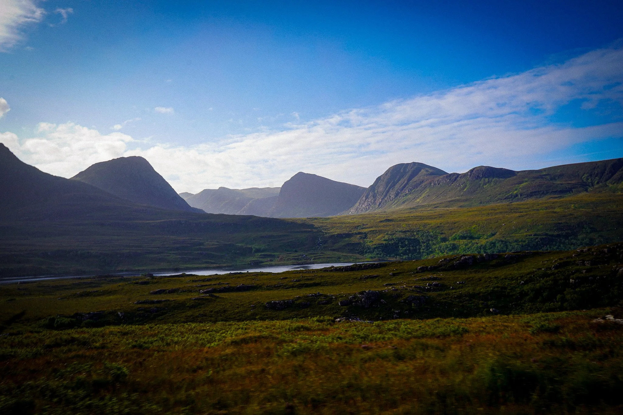 A scenic landscape with rolling green hills, a lake, and distant mountains under a partly cloudy blue sky.