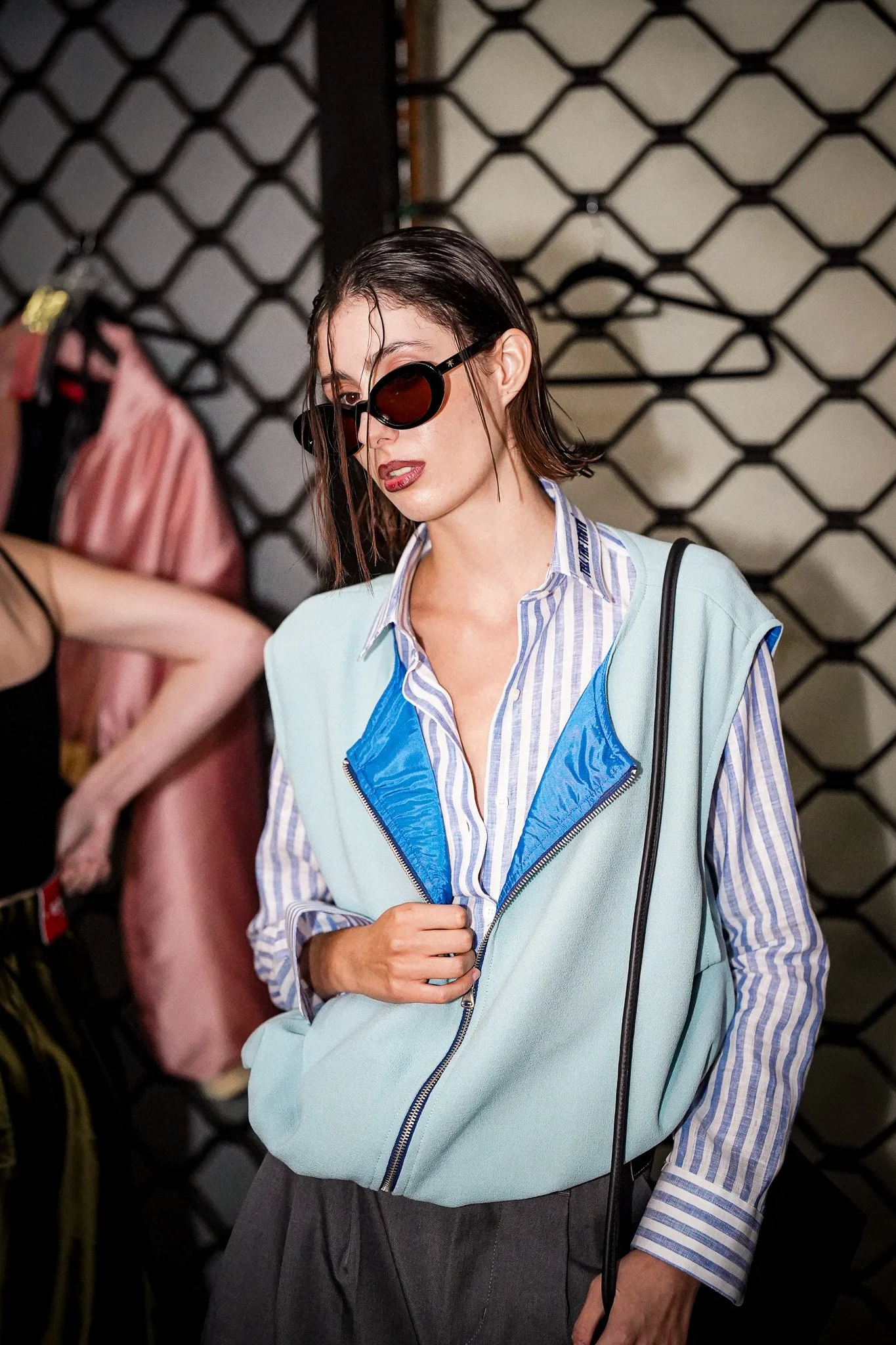 A woman with wet hair wearing large black sunglasses, a blue and white striped shirt, and a light blue sleeveless zip-up jacket. She is holding her jacket near her waist with her right hand, and a black strap of a bag is over her left shoulder. She i