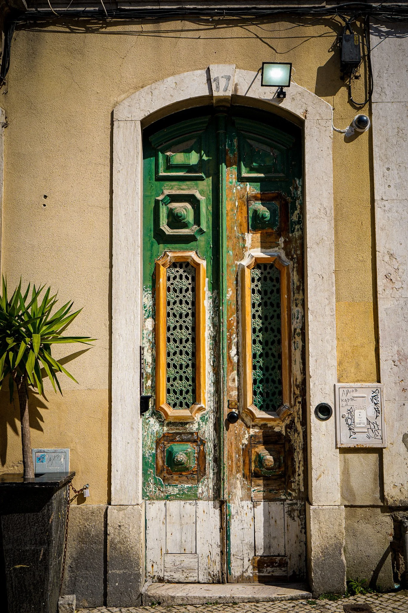 An old, weathered green and white double door with intricate cutout designs, set in a stone and stucco building. The door shows signs of peeling paint and age, and is framed by a stone arch with the number 17 above it. There are security cameras and 