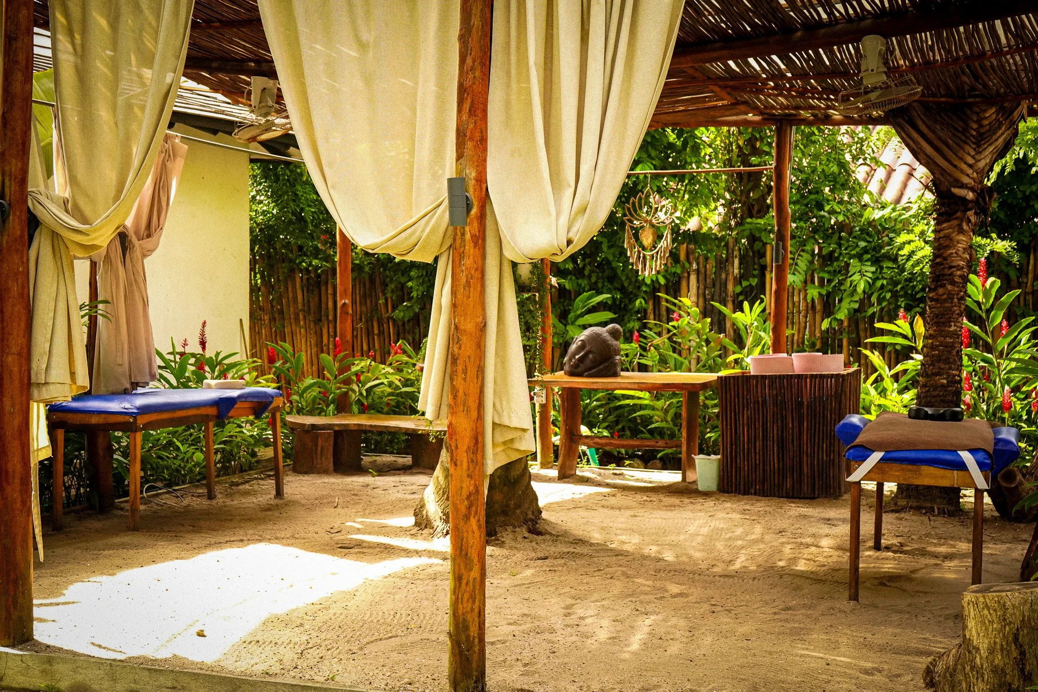 Outdoor shaded seating area with wooden furniture, curtains, tropical plants, and decorative hanging art.