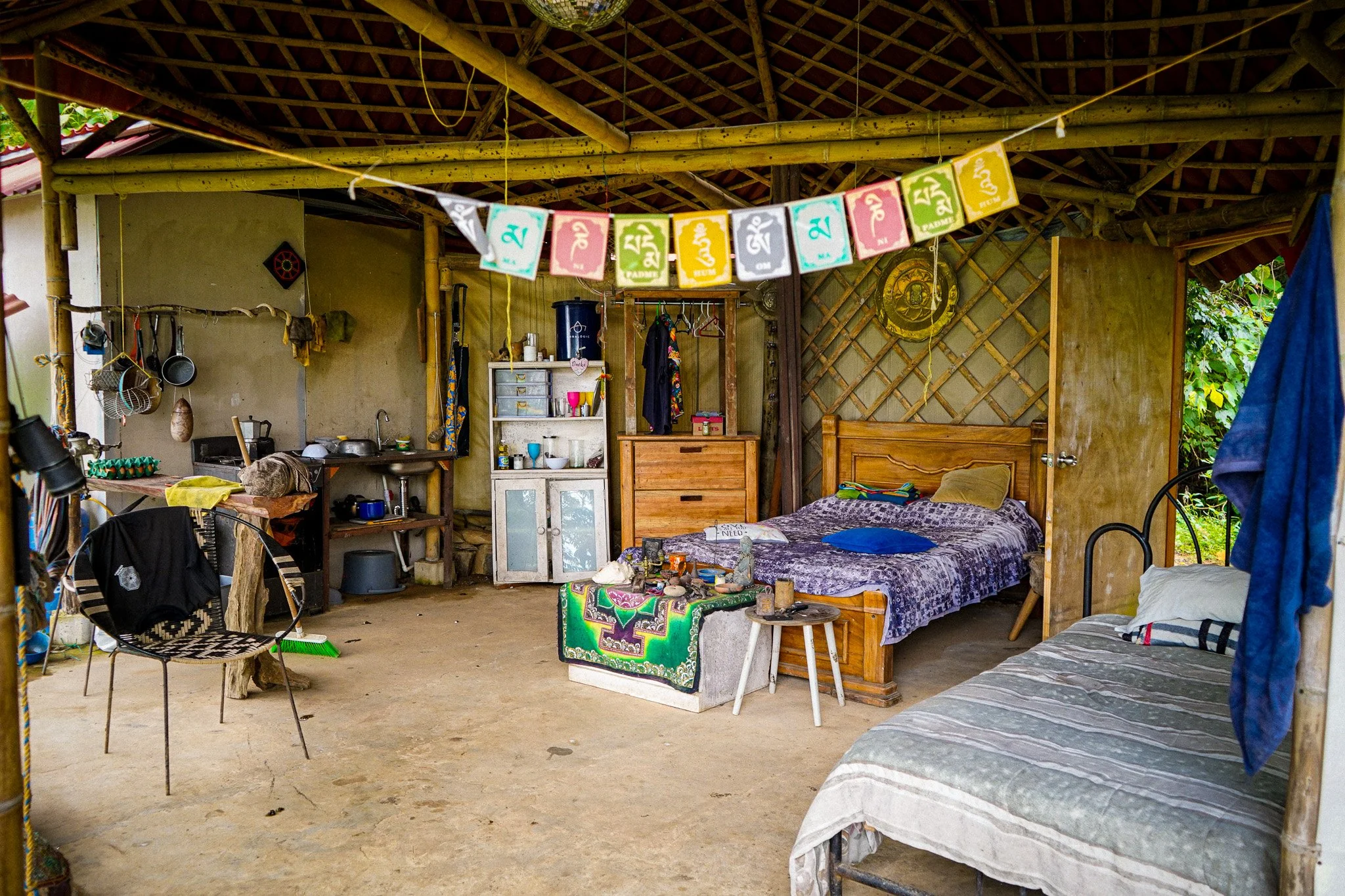 Interior of a rustic bamboo and thatch bungalow with a bed, a small table, kitchen essentials, and colorful banners hanging from the ceiling.