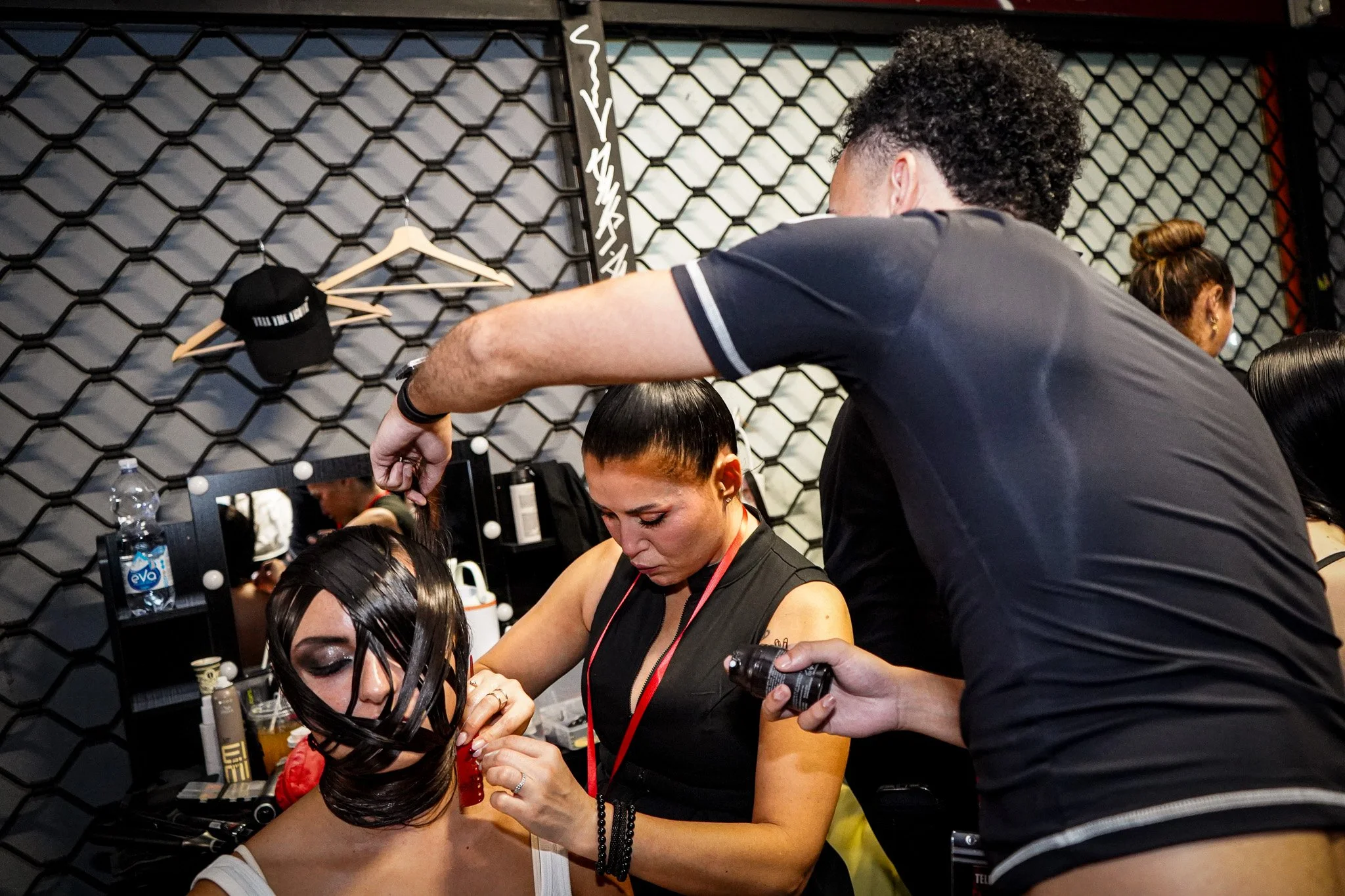 A woman getting her hair styled at a beauty salon, with a stylist working on her hair while holding a styling product. The background features a black hexagonal grid wall with various items, a water bottle, and a black cap hanging on the wall.