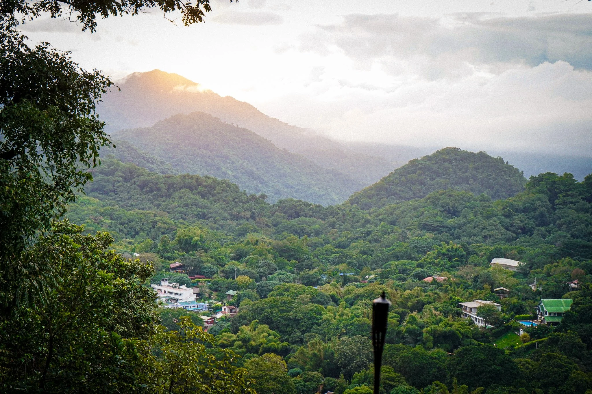 Scenic view of a lush green mountainous landscape with trees and a few buildings at the base, partly cloudy sky with sunlight breaking through.