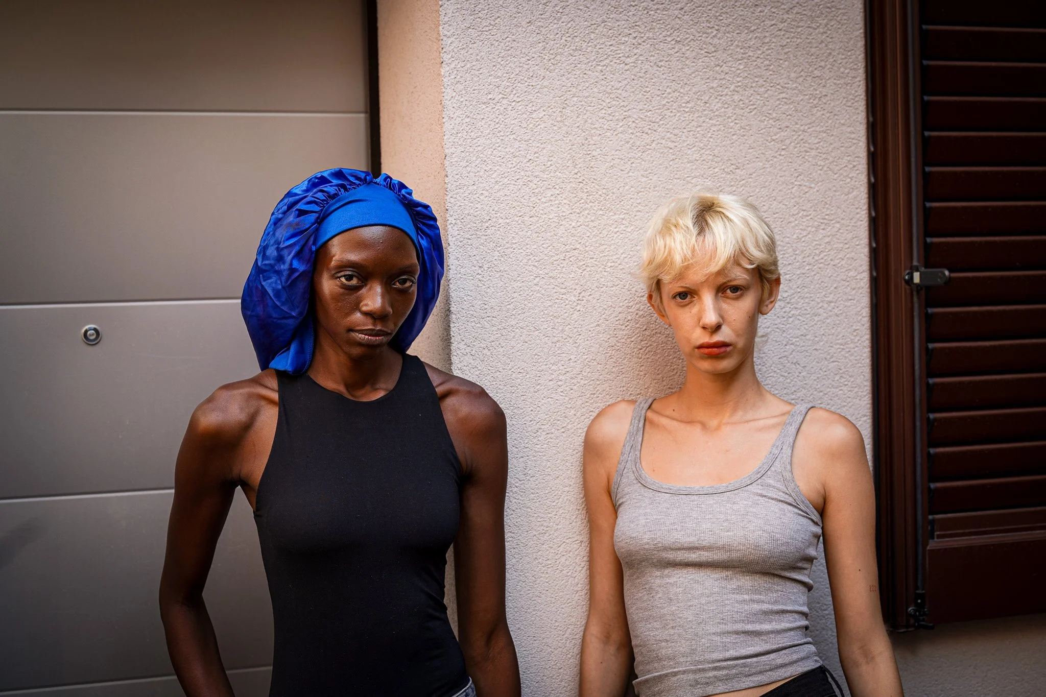 Two models. standing side by side against a wall outside, one with dark skin and blue headwrap and the other with light skin and short blonde hair, both wearing tank tops.