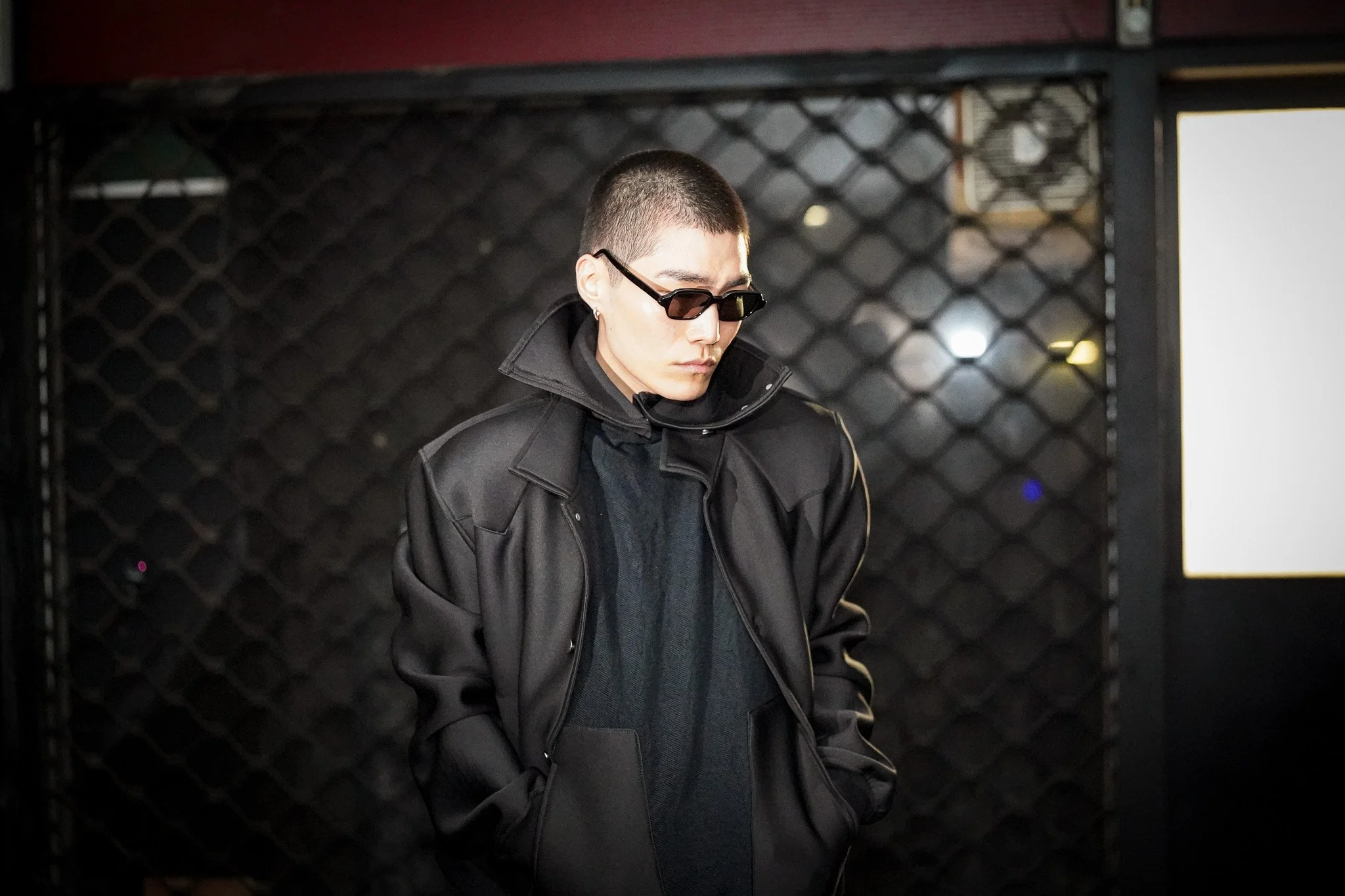 A person with short hair and sunglasses wearing a black jacket, standing in front of a black, diamond-patterned background.