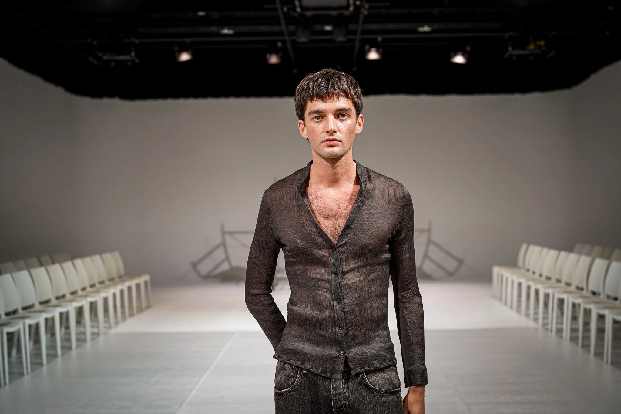 A model man with dark hair and a serious expression, wearing a semi-transparent gray shirt, standing in a room with white chairs arranged in rows on either side. The ceiling has visible rigging and lighting equipment.