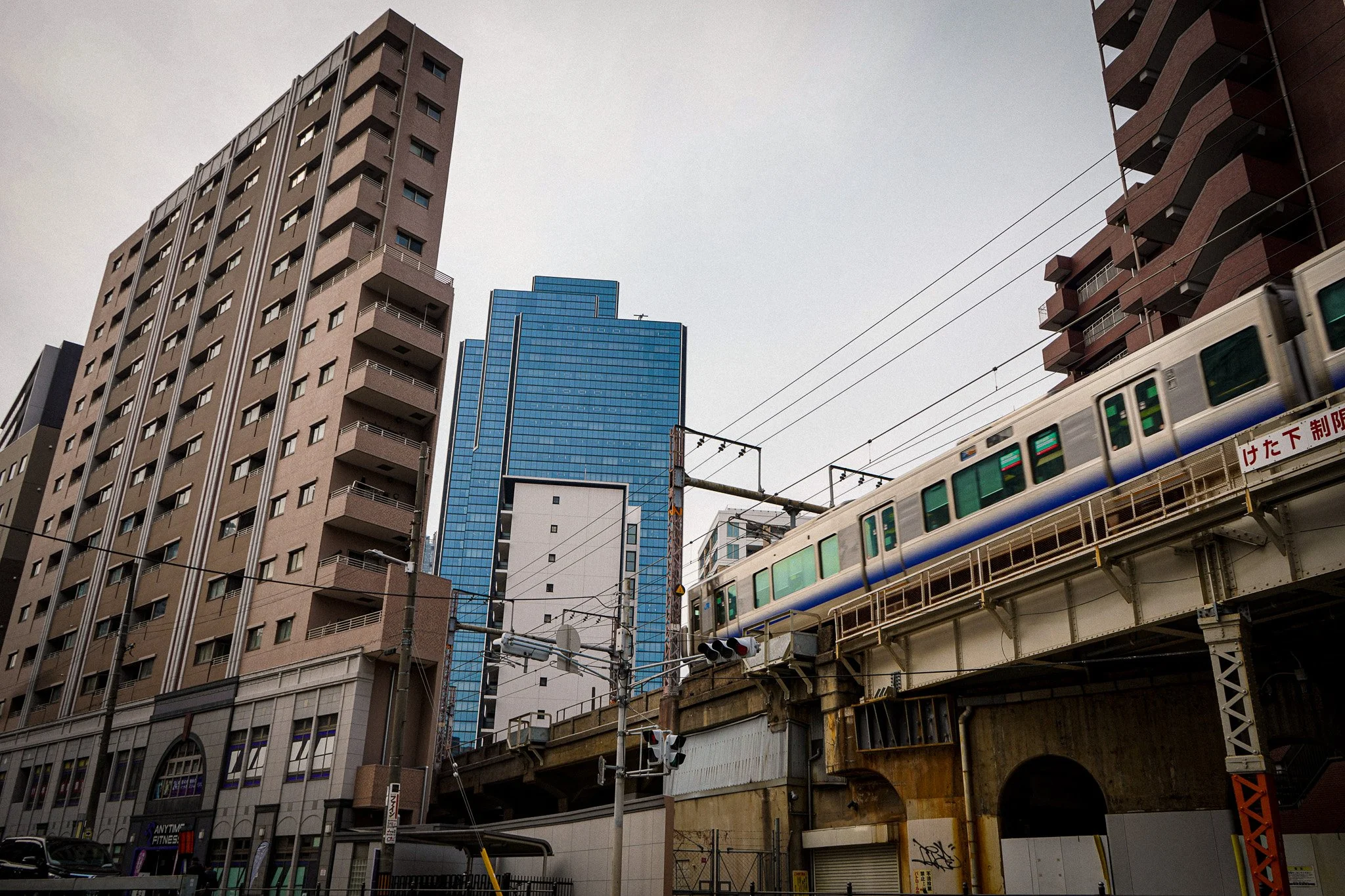 Cityscape with tall buildings, a train on elevated tracks, and wires overhead, under a gray sky.