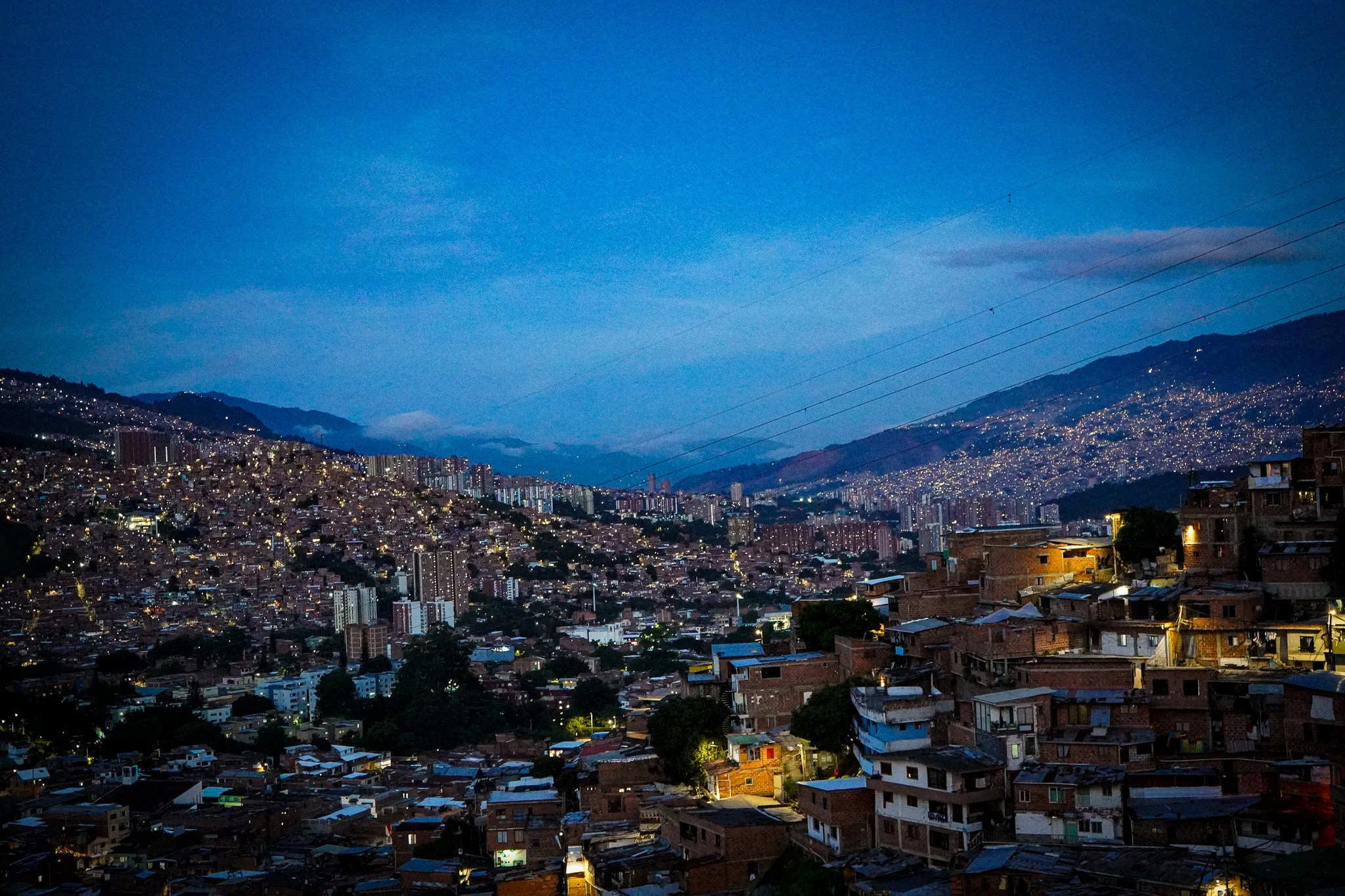 A cityscape at dusk with numerous buildings and houses covering the hills, illuminated by streetlights with a darkening blue sky overhead.