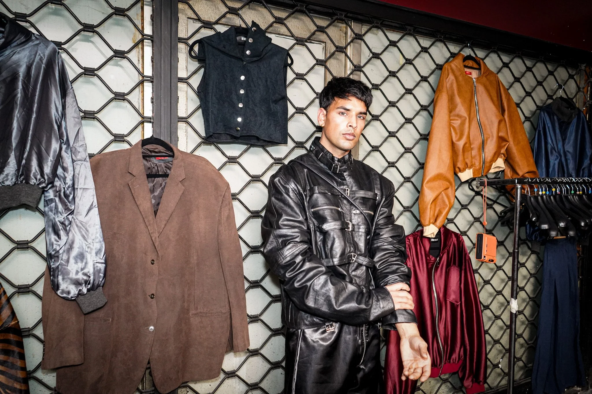 A young man with dark hair and light skin, wearing a black leather jacket, standing in a clothing store. Behind him is a display wall with various jackets and clothing items hanging, including a brown blazer, a black sleeveless jacket, a tan bomber j