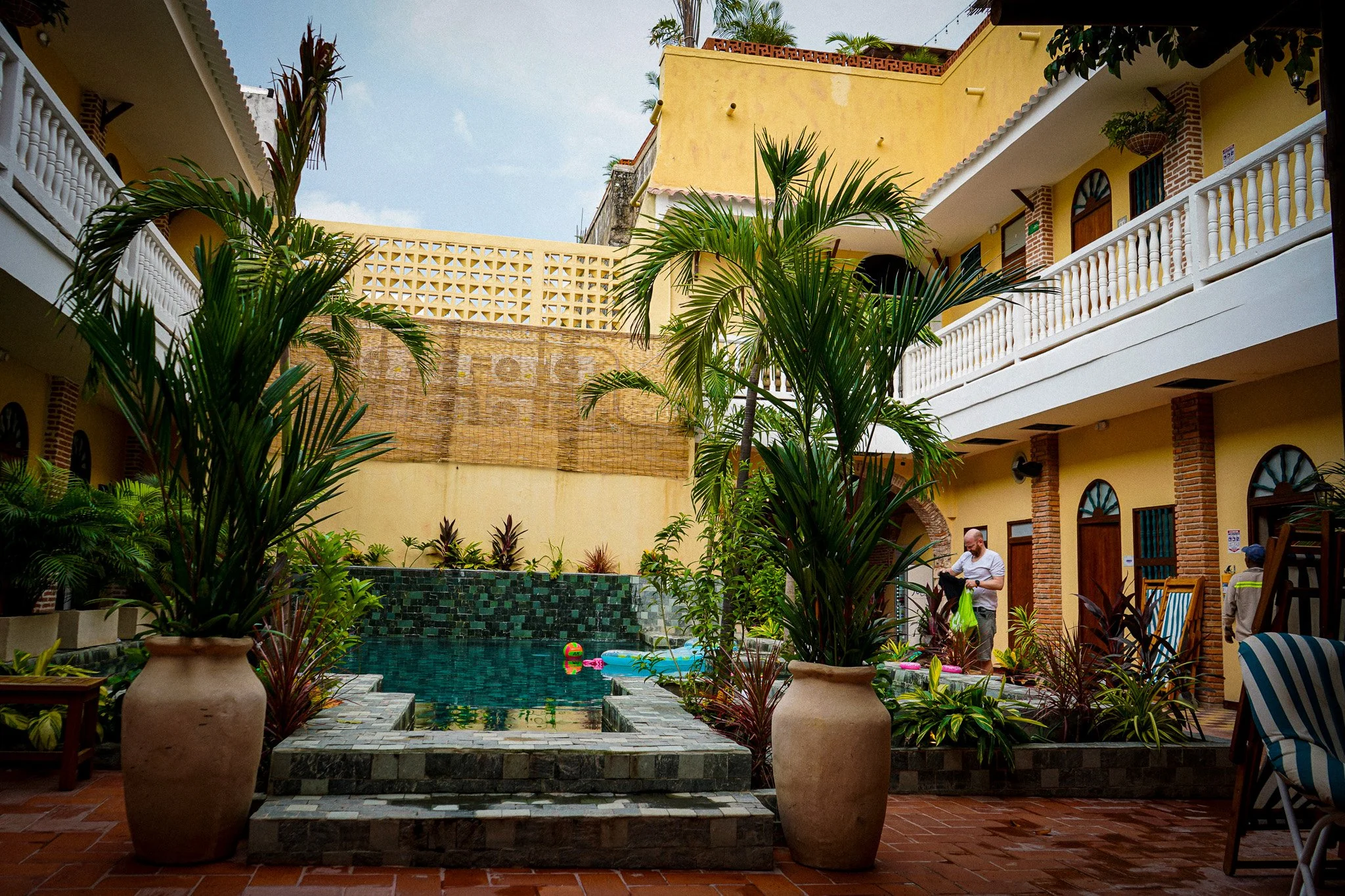 A courtyard with a swimming pool surrounded by plants, potted trees, and outdoor furniture, with multi-story yellow and brick buildings in the background.