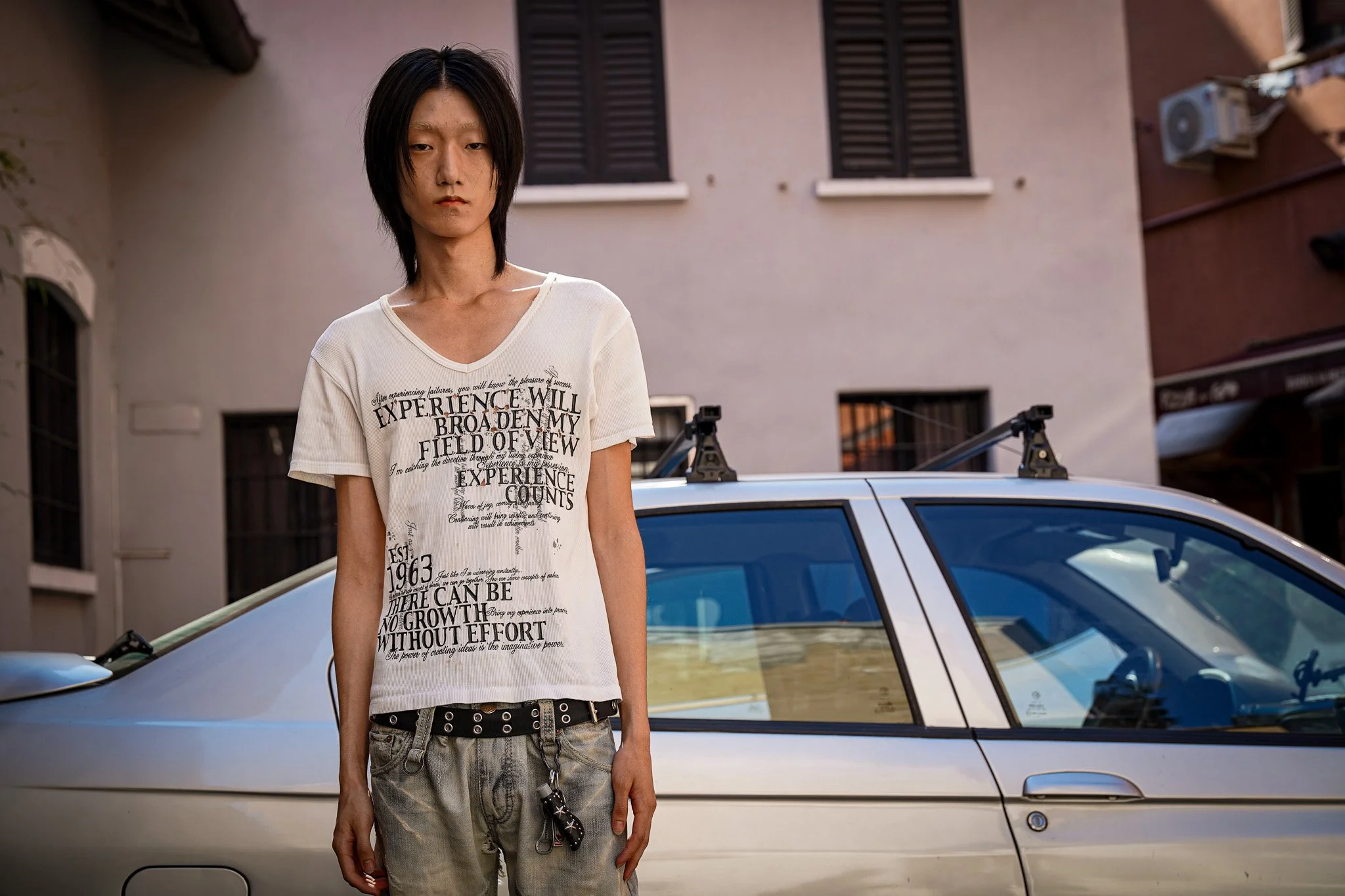A young asian male with straight black hair and a serious expression standing in front of a silver car, wearing a white graphic T-shirt with text and gray jeans, in an urban residential area with buildings and closed windows in the background.