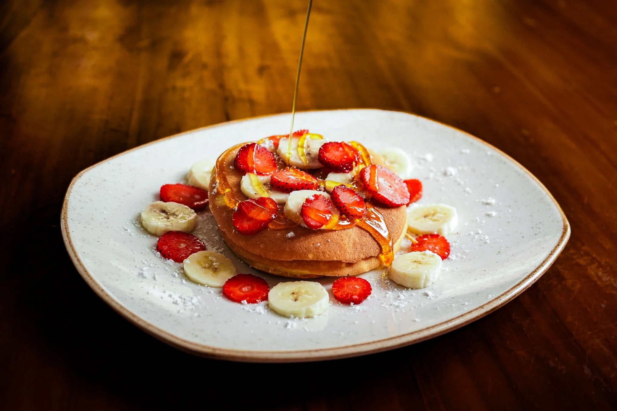Close-up of a plate with a waffle topped with sliced strawberries and bananas, drizzled with syrup, and dusted with powdered sugar, with syrup being poured over it.