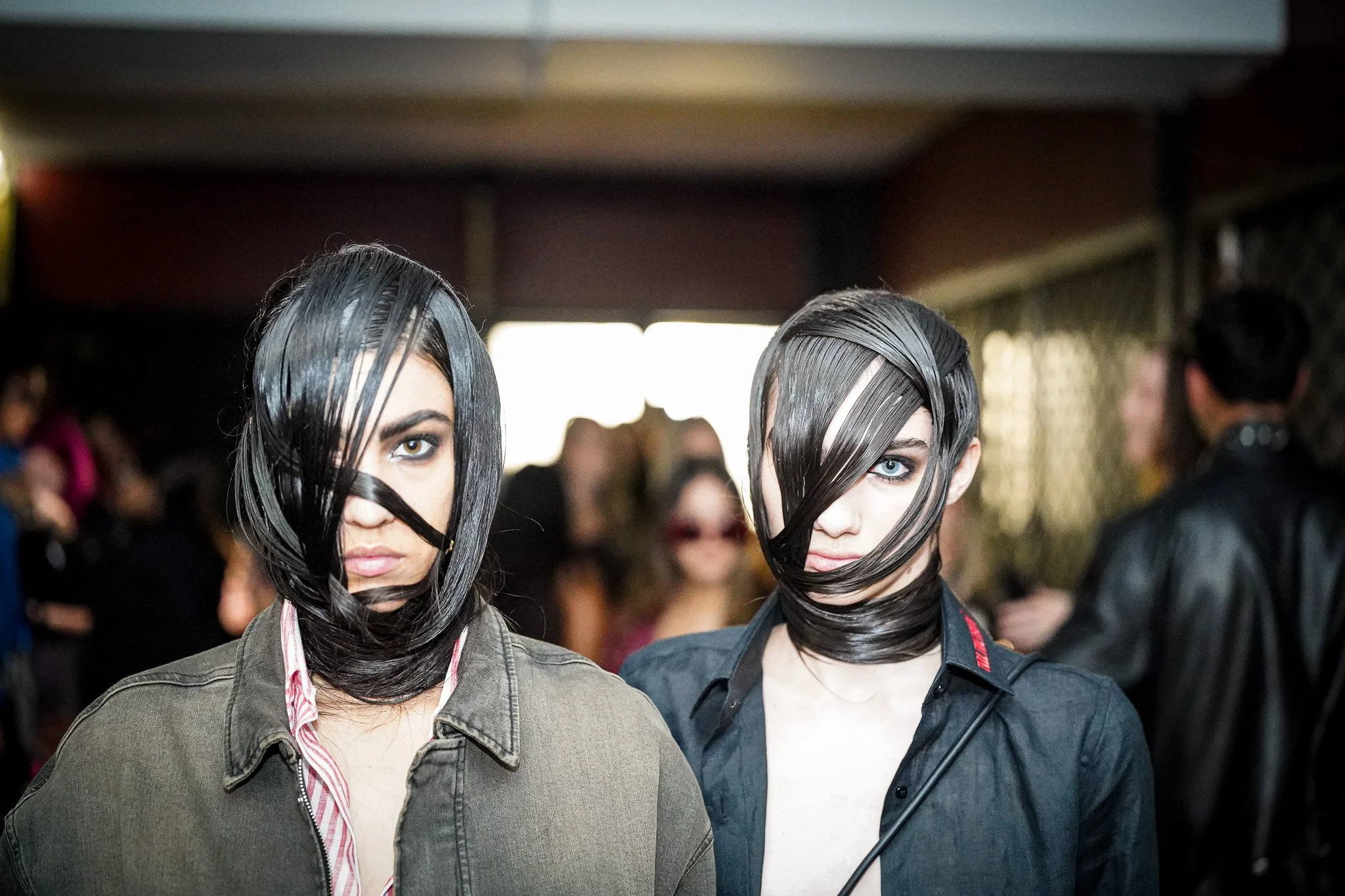 Two women with hair styled in a creative fashion, with strands wrapped around their faces, standing in a crowded indoor setting.