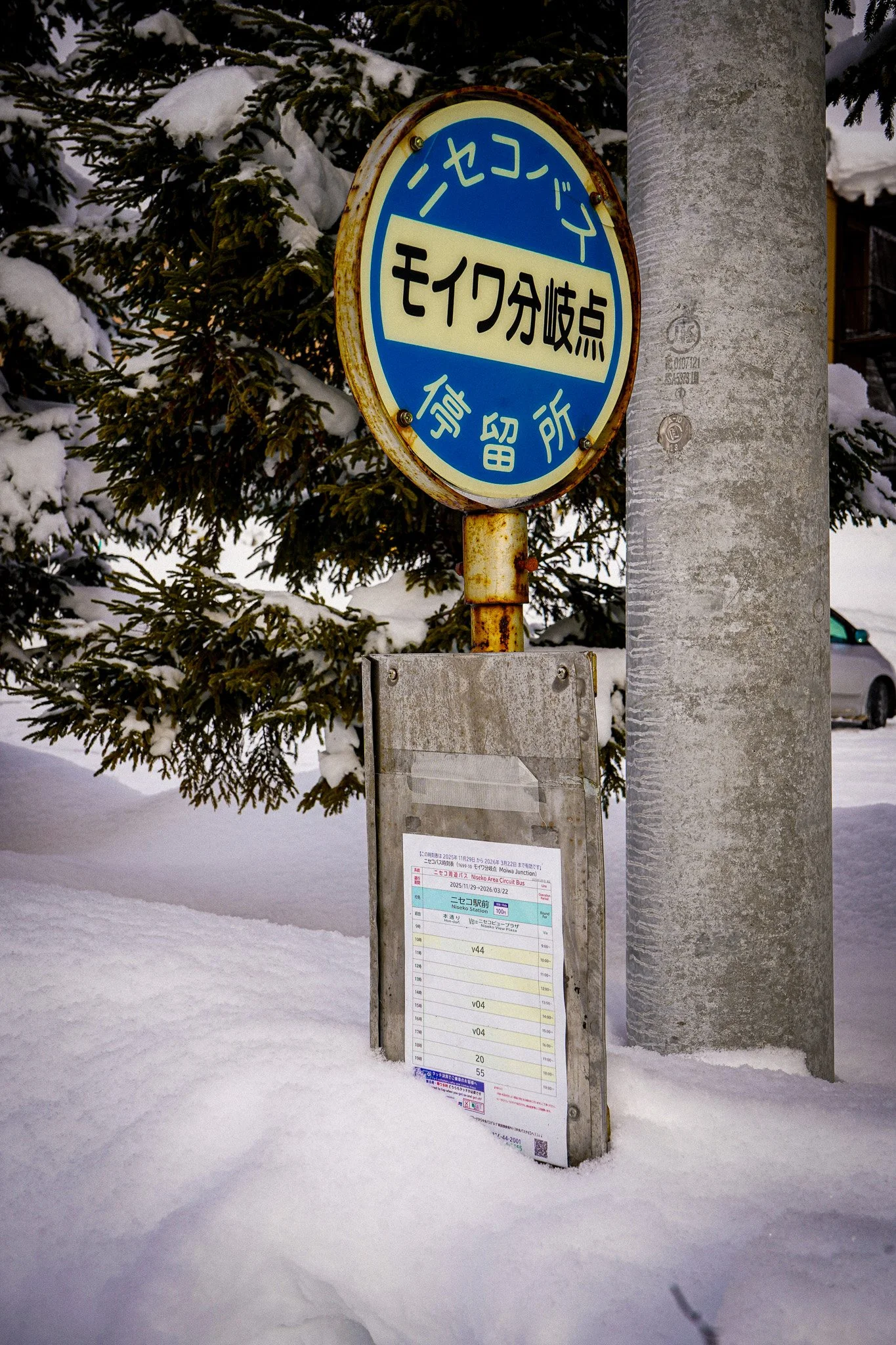 A blue and yellow bus stop sign with Japanese writing, mounted on a pole in a snowy outdoor setting, with snow-covered trees and a building in the background.