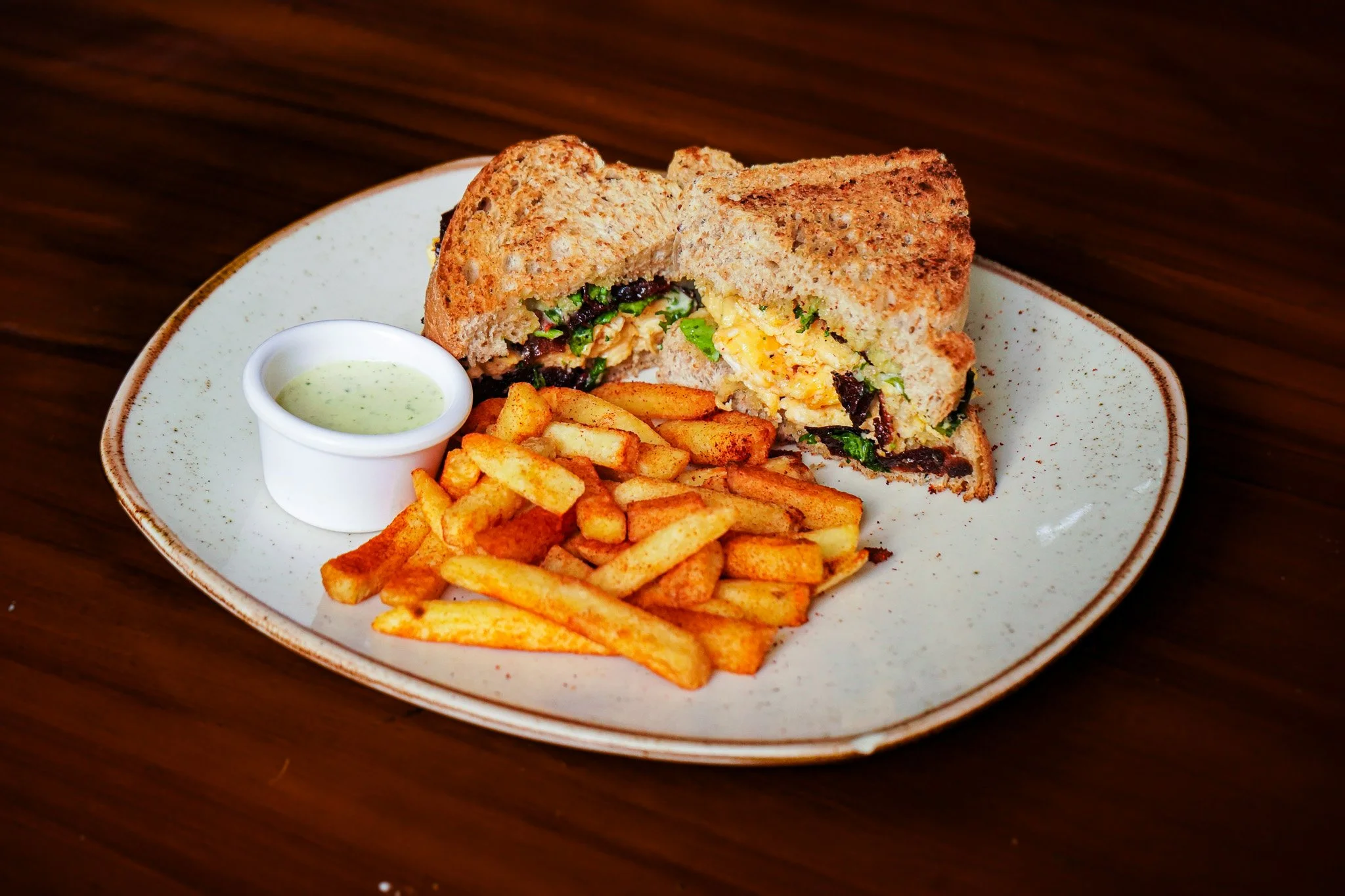 A plate with a sandwich filled with scrambled eggs, black beans, and greens, French fries, a small container of green dipping sauce, on a wooden table.
