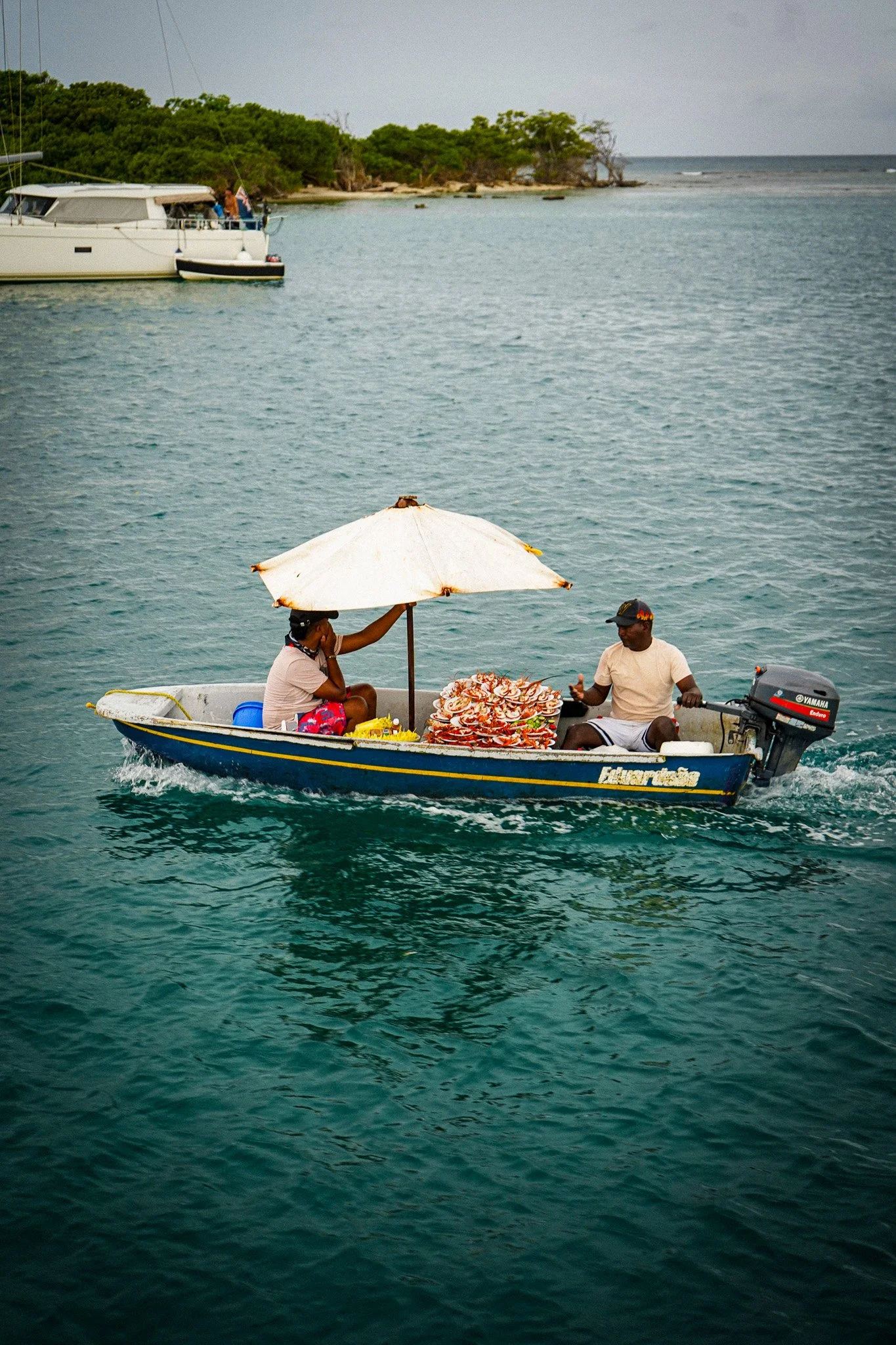 A small boat with two people selling seafood is moving through the water near a rocky and green shoreline. One person is sitting under a white umbrella, and the other is standing at the outboard motor, which is labeled Yamaha.