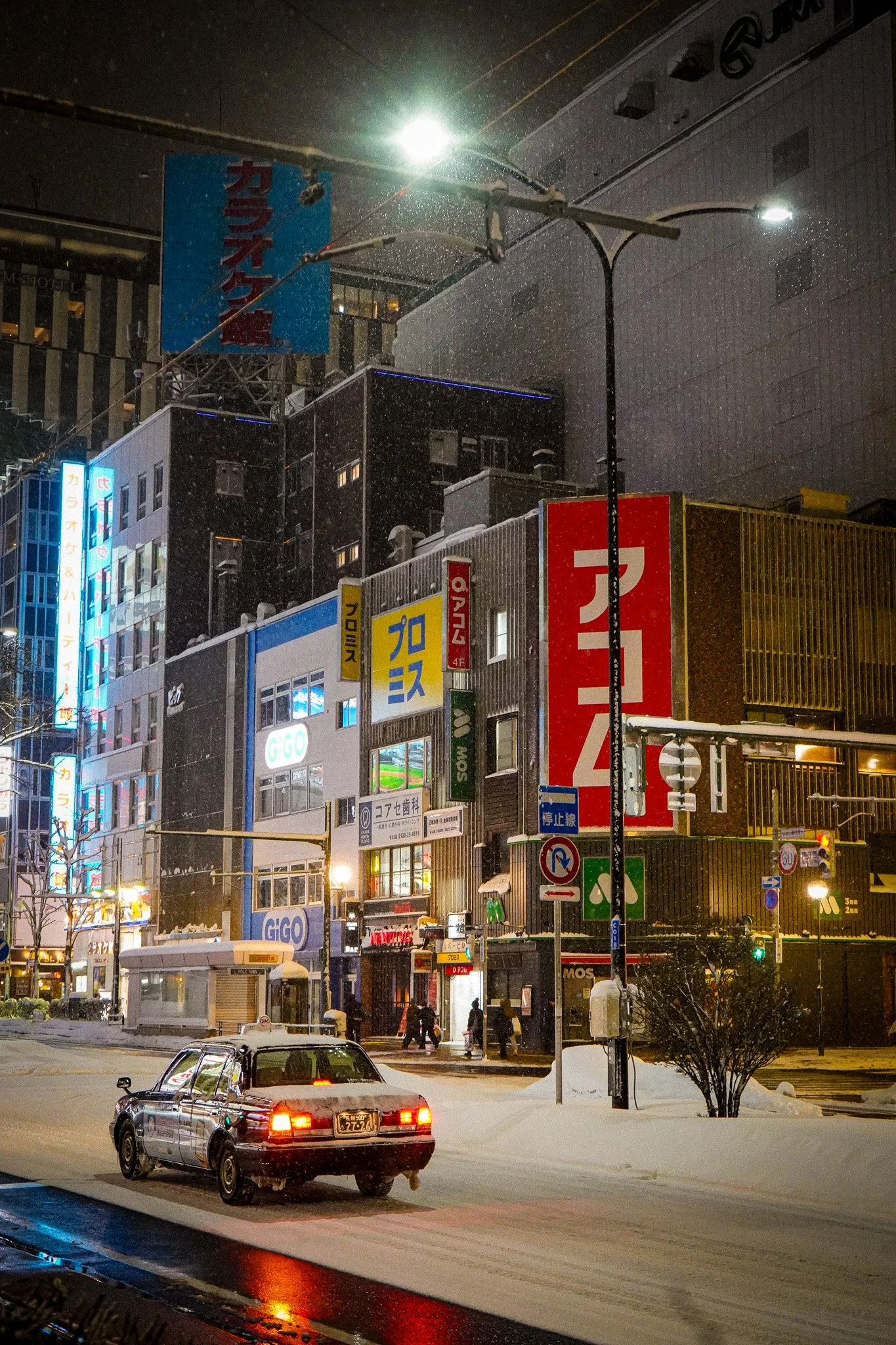 Nighttime scene of a snowy city street with a dark sky, streetlights, and illuminated signs on buildings. A black car is parked on the snow-covered road, and pedestrians are walking on the sidewalk.