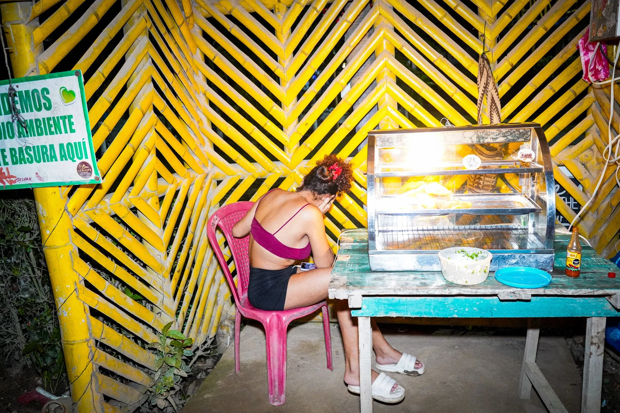 A woman with curly hair, wearing a sleeveless purple top and white sandals, sitting at a rustic table in front of a brightly painted yellow and black geometric patterned wall. She appears to be covering her face with her hand. There is a food display