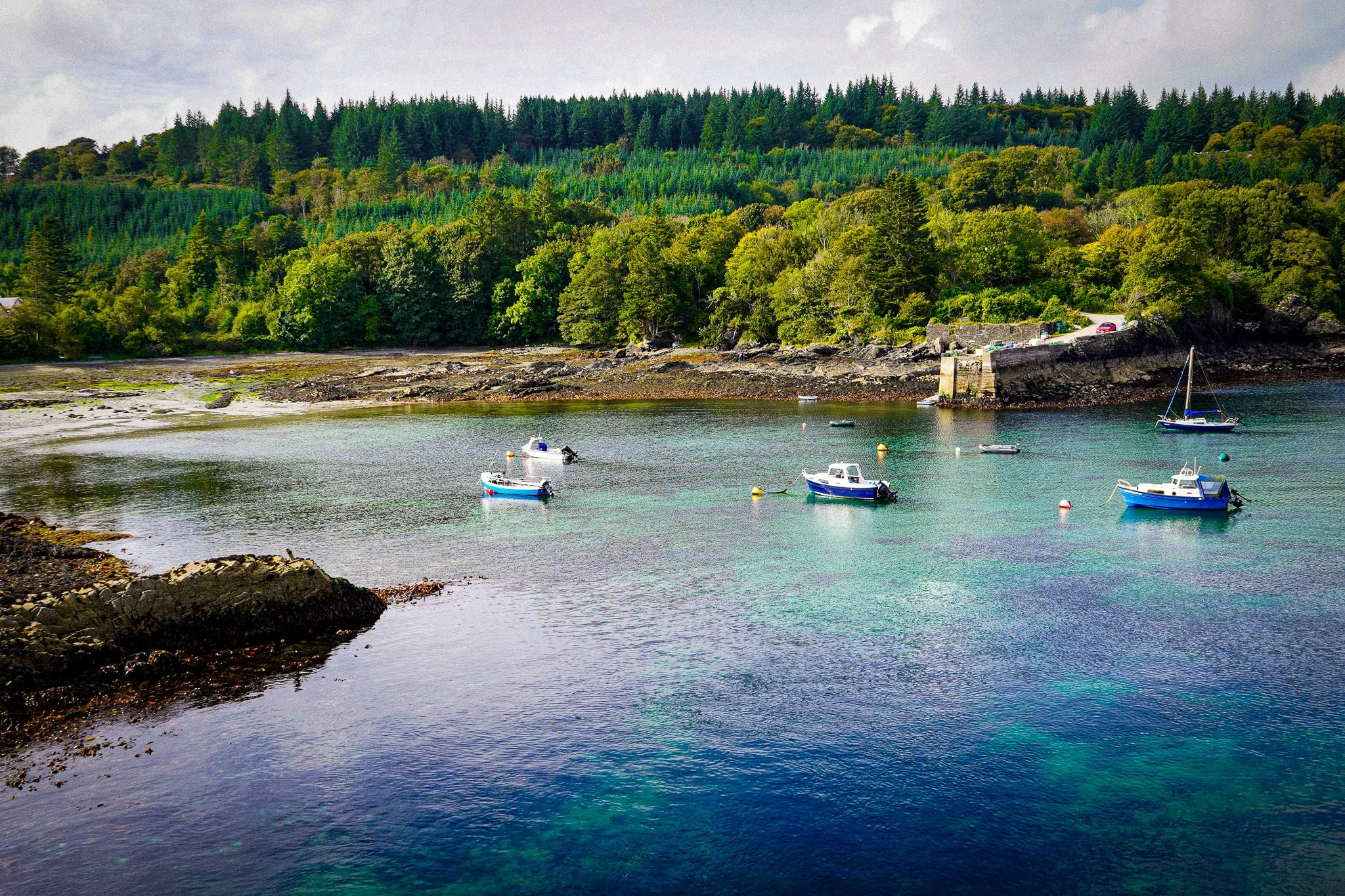 A coastal scene with boats floating on calm water, green forested hills in the background, rocky shoreline in the foreground, and cloudy sky overhead.