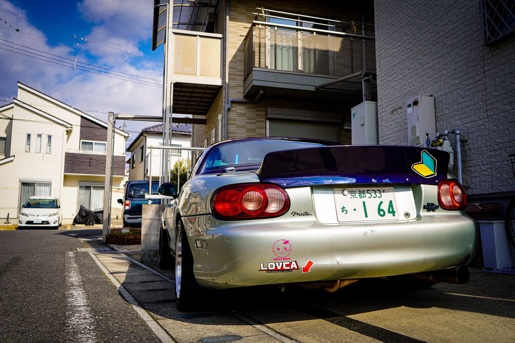 A silver sports car with stickers and Japanese license plates parked in a residential parking lot, with multi-story houses and a blue sky in the background.